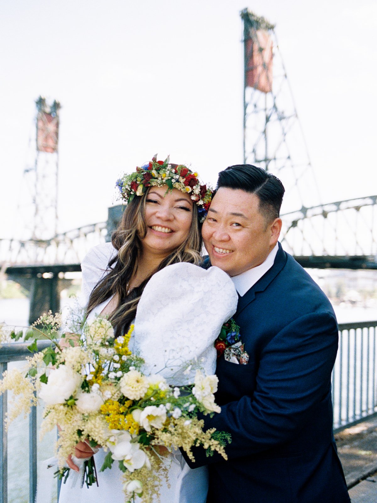 Smiling newlyweds pose together in front of a steel bridge, with the bride holding a loose white and yellow bouquet.