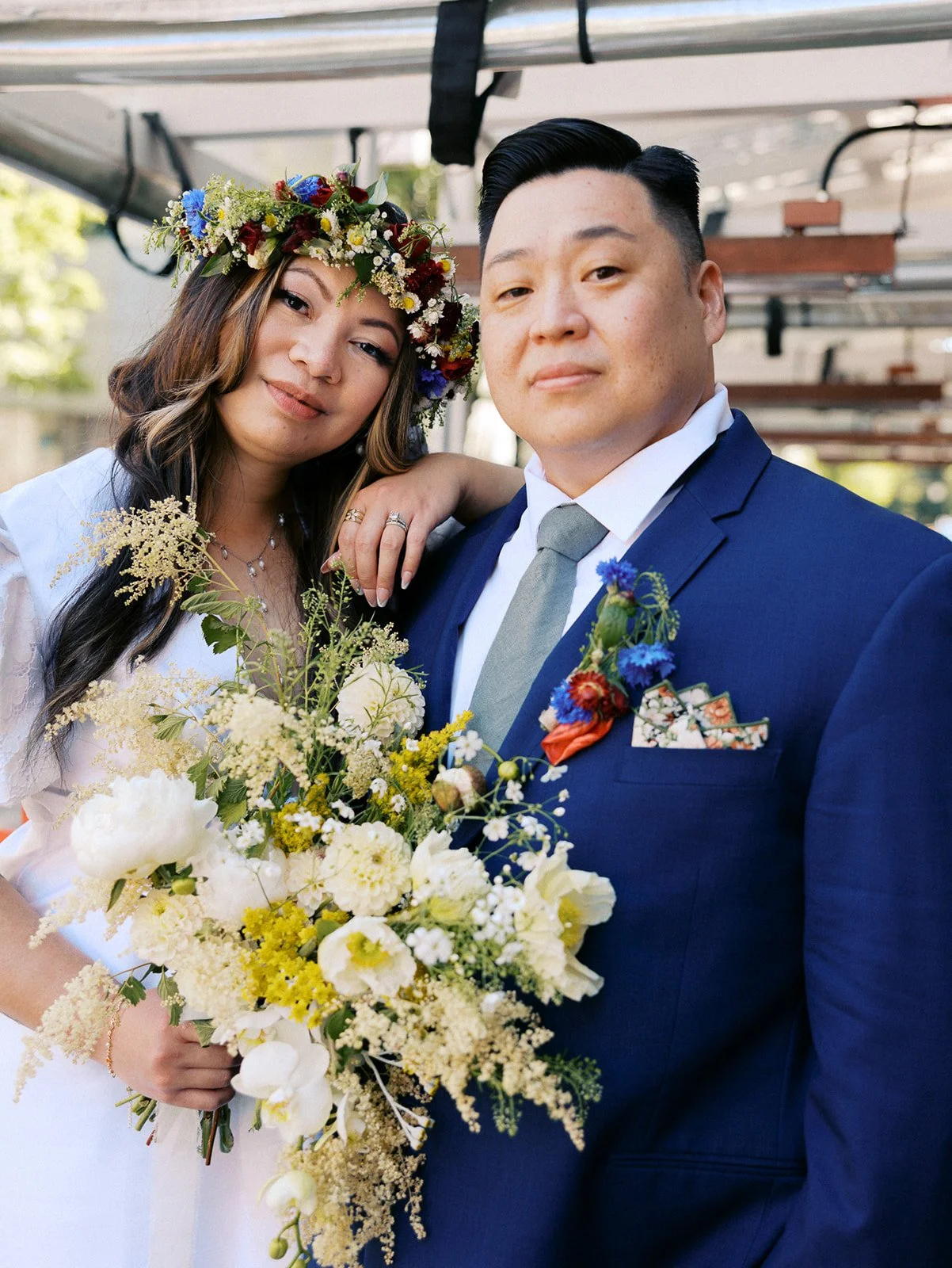 Close-up of a couple posing together, featuring the bride's wildflower crown, white bouquet, and groom's navy blue suit.