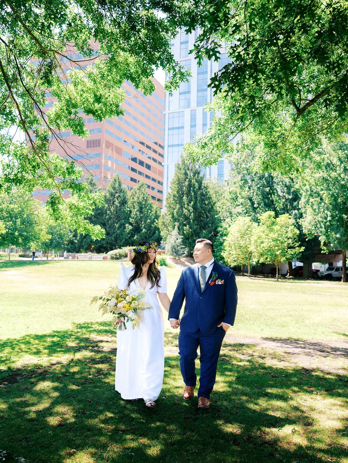 Newlyweds walk hand in hand through a sunny downtown Portland park surrounded by green trees and city buildings.
