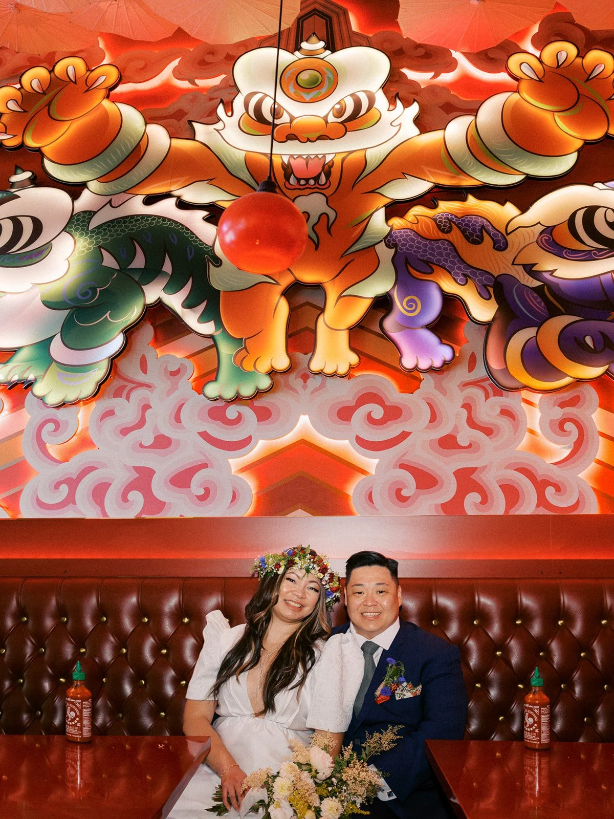 Newlyweds sit together in a red leather restaurant booth beneath a colorful lion dance mural after their wedding.