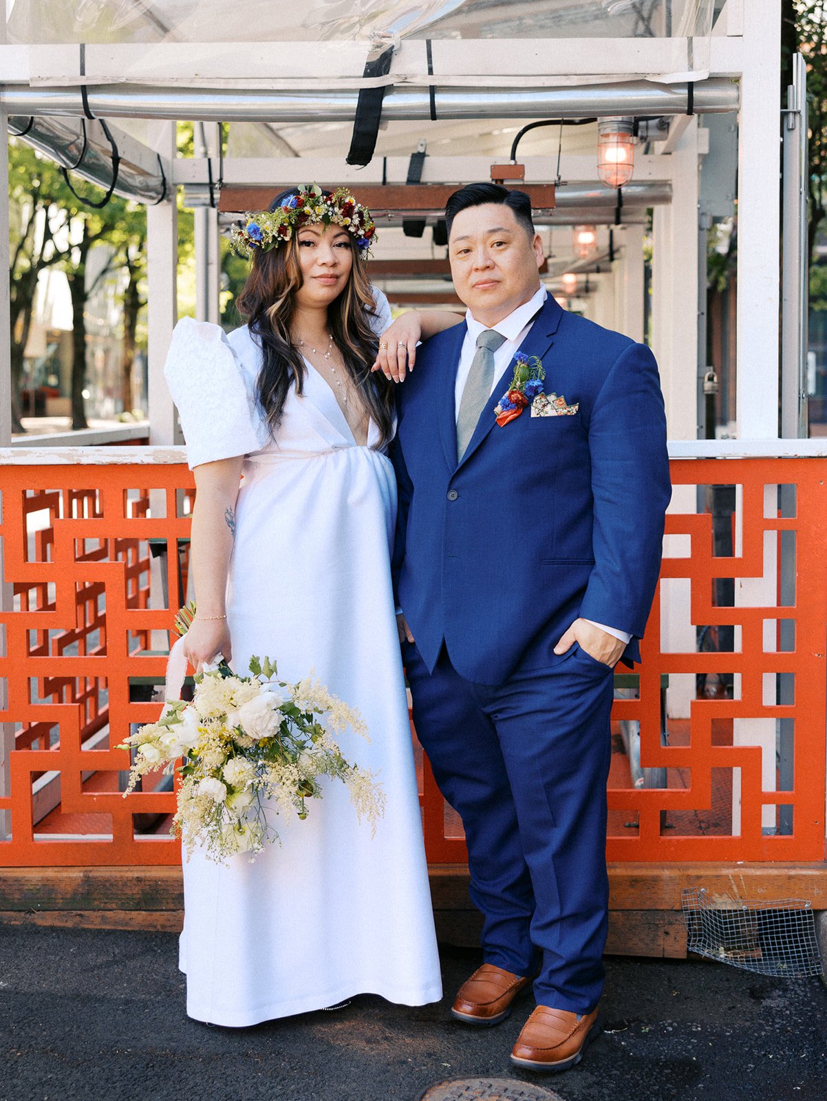 Newlyweds pose in front of a bright red geometric railing, with the bride holding a loose bouquet of white and yellow flowers.