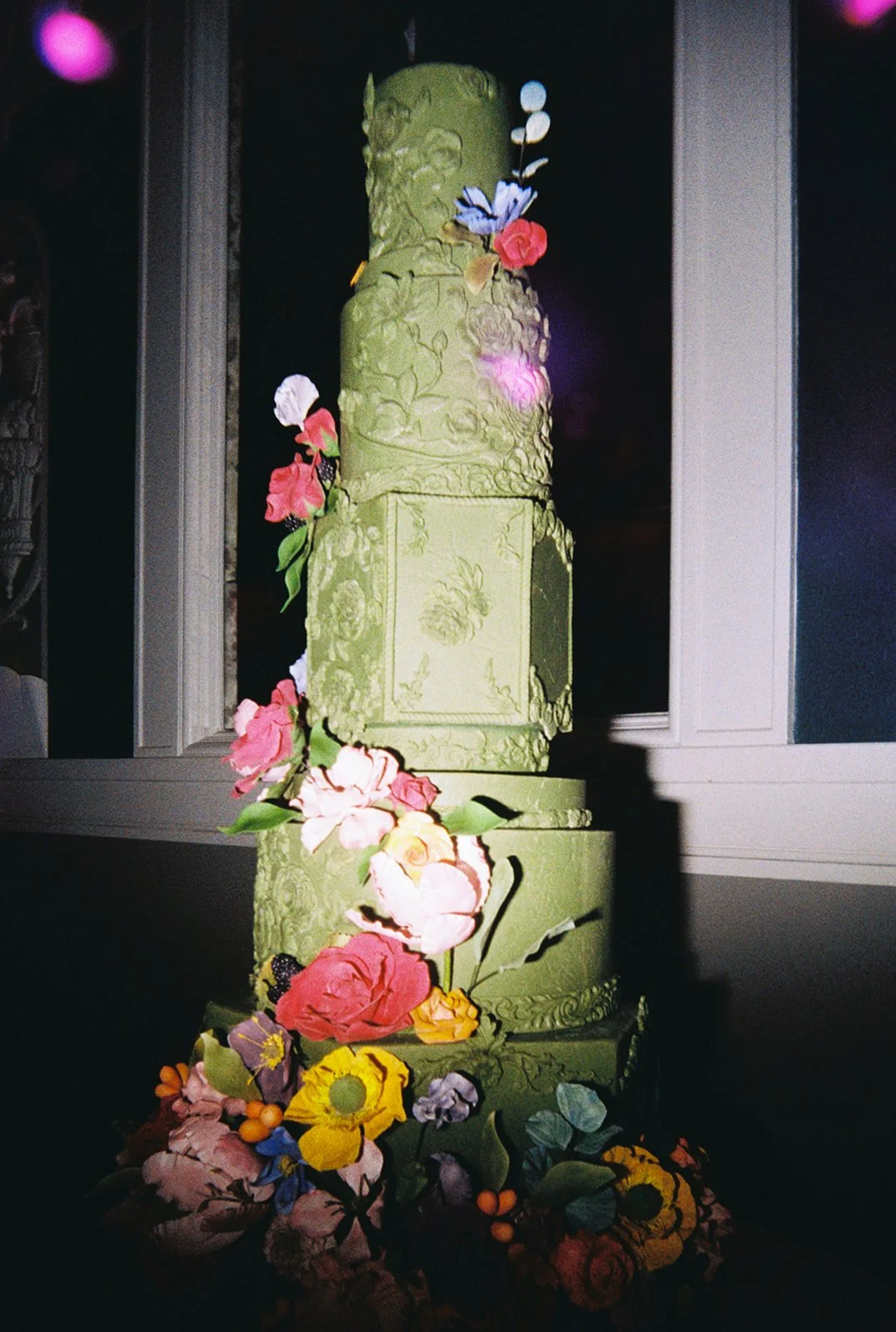 Multi-tier green wedding cake with floral decorations and textured icing displayed in dim lighting.