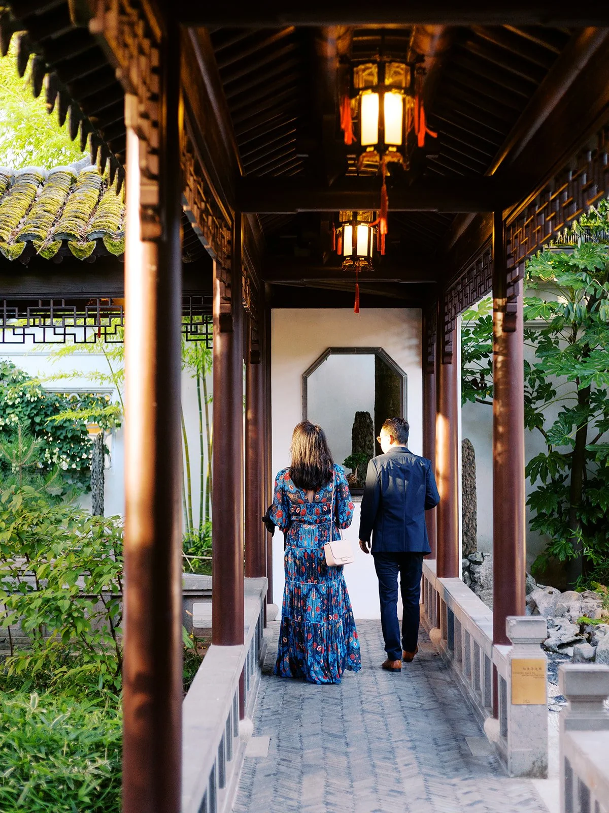 Two guests — a woman in a floral maxi dress and a man in a navy suit — walk along a covered gallery corridor hung with glowing red lanterns.