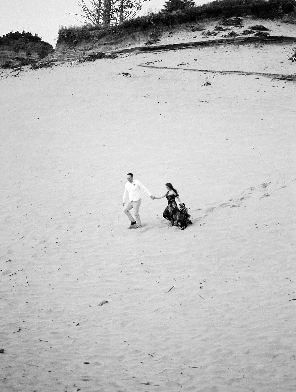 Black and white image of couple walking across sandy Oregon dunes, holding hands from a distance