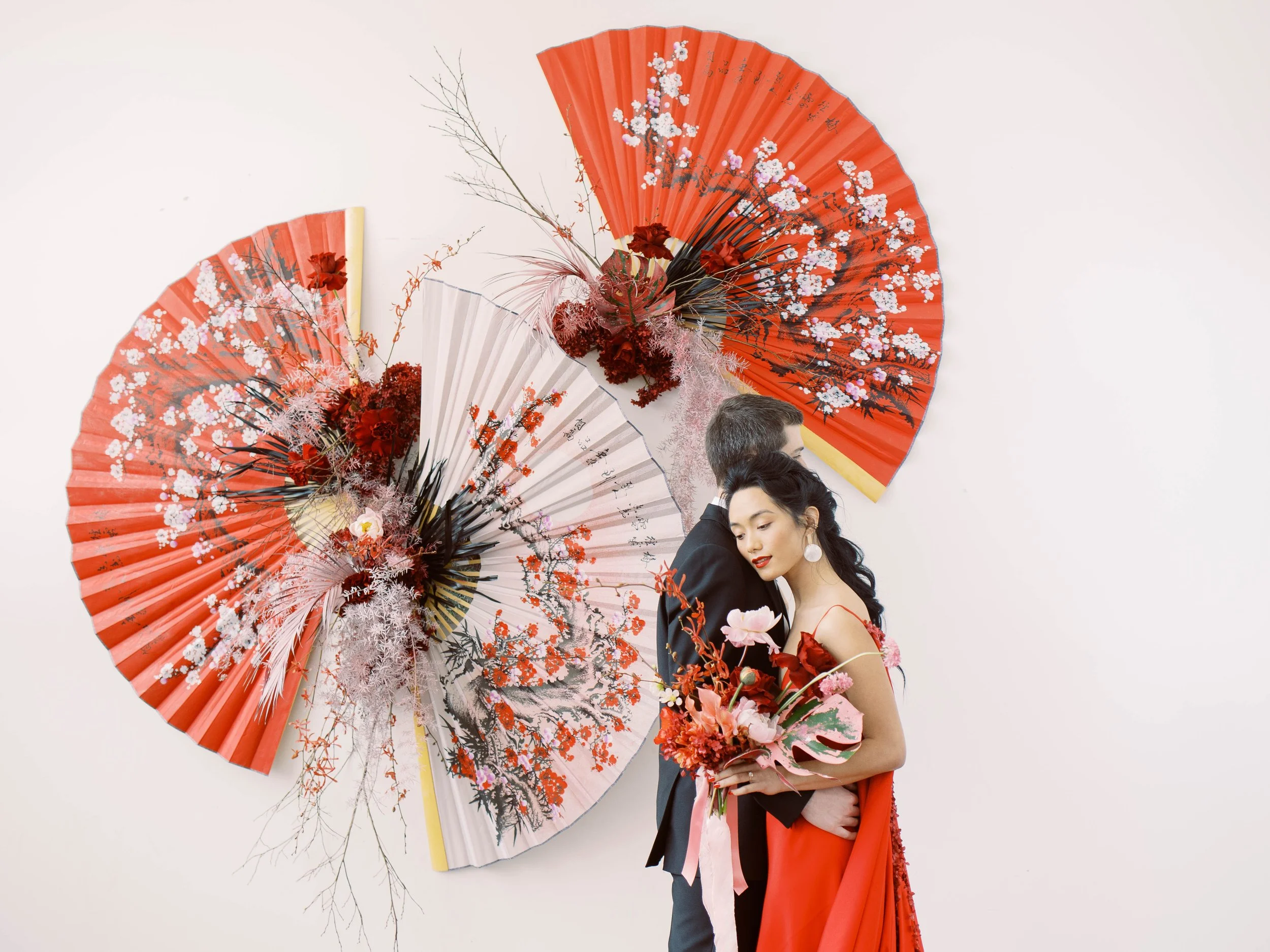 Couple poses in formalwear beside oversized red and white Chinese fans decorated with flowers.