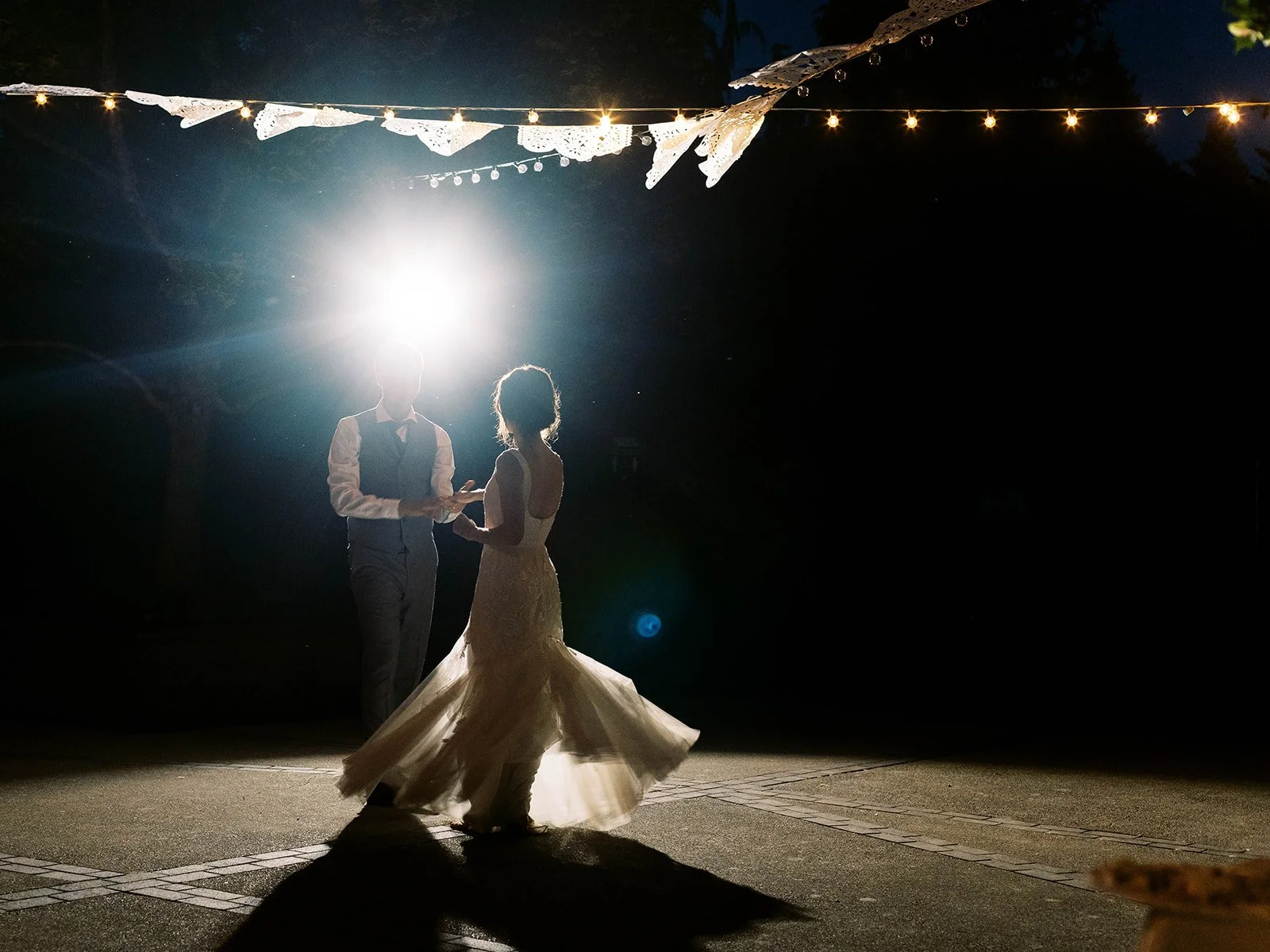 Bride and groom sharing first dance at night under string lights with dramatic backlighting