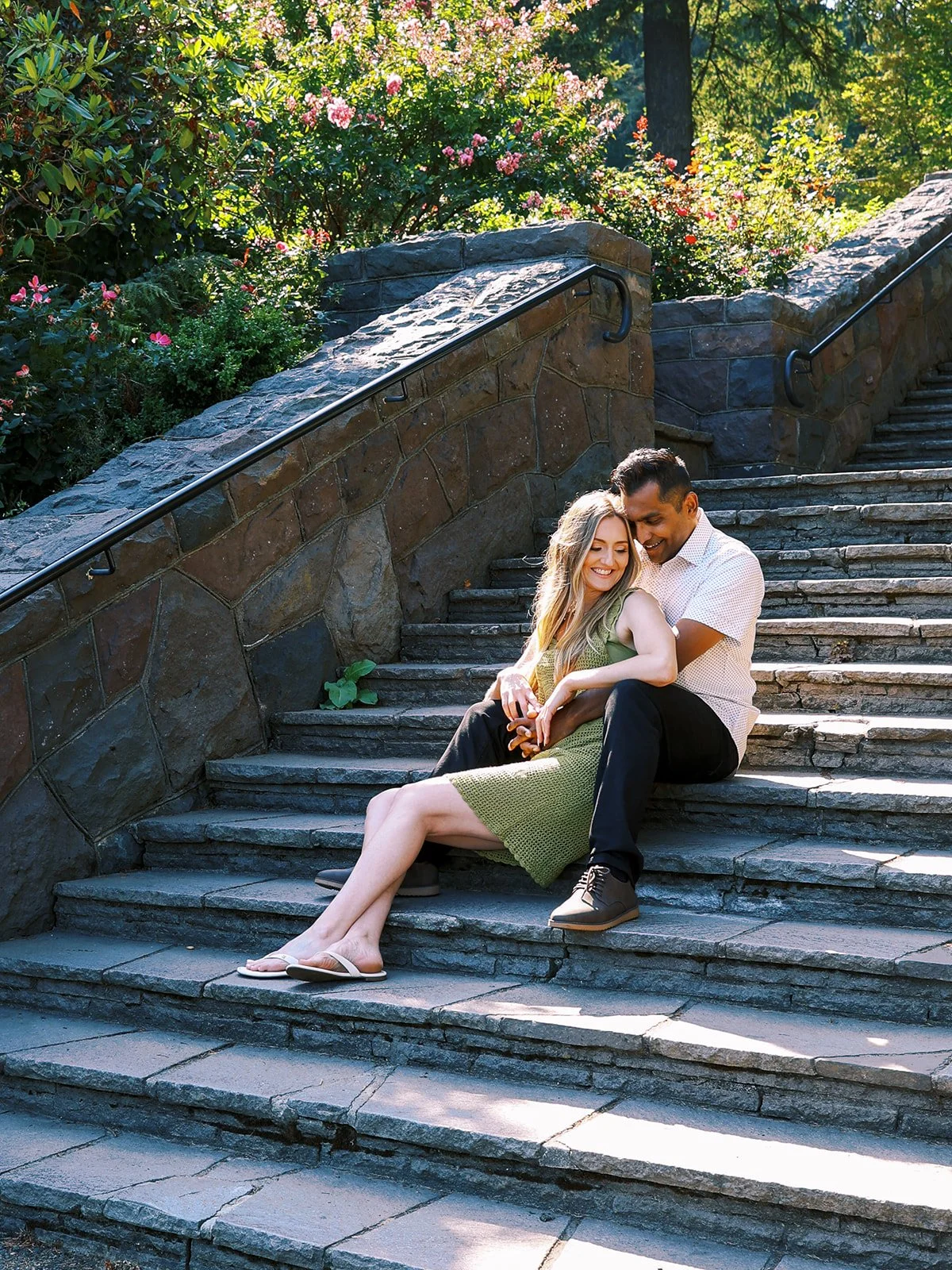 A couple cuddled together on stone garden steps, surrounded by blooming pink roses and greenery.