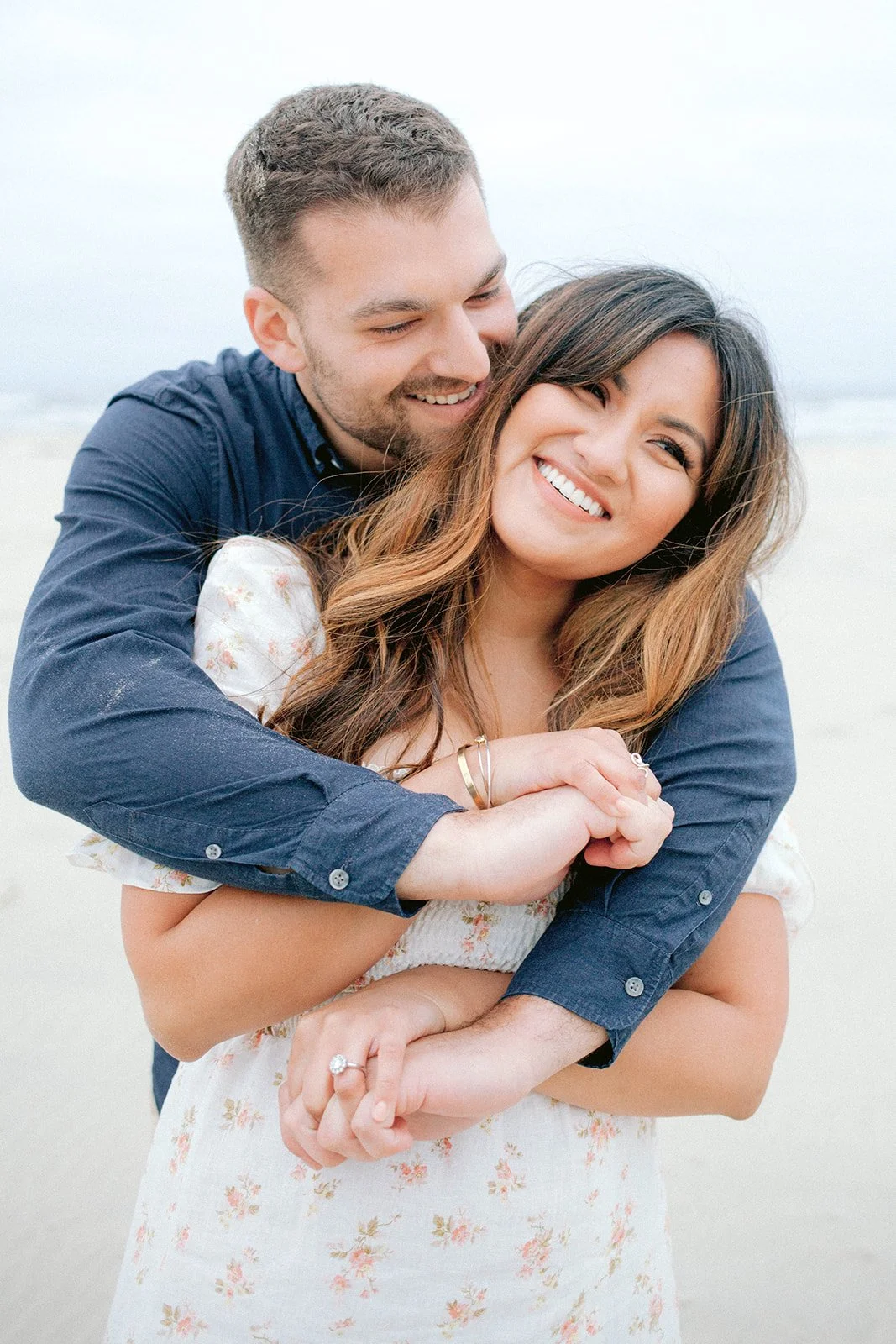 Man hugging woman from behind on beach, both smiling in soft natural coastal light
