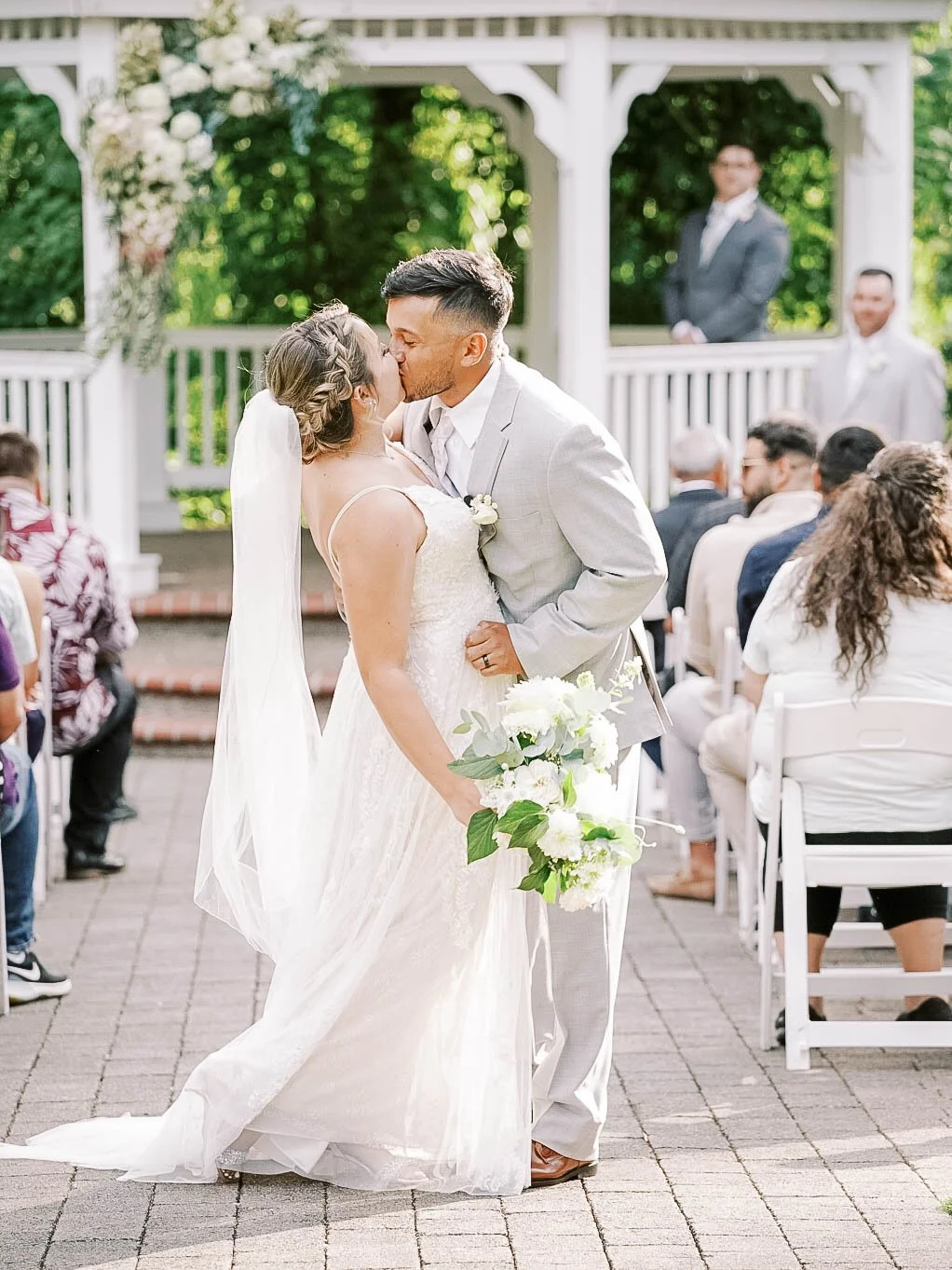 Newlywed couple shares a kiss in the aisle after the ceremony, bride holding a white bouquet as guests watch from both sides.