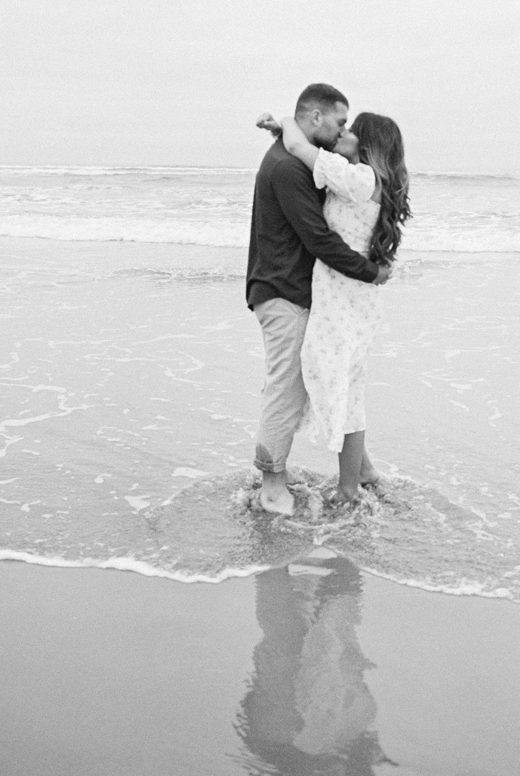 Couple kissing while standing in shoreline waves, reflected in wet sand, black and white photo