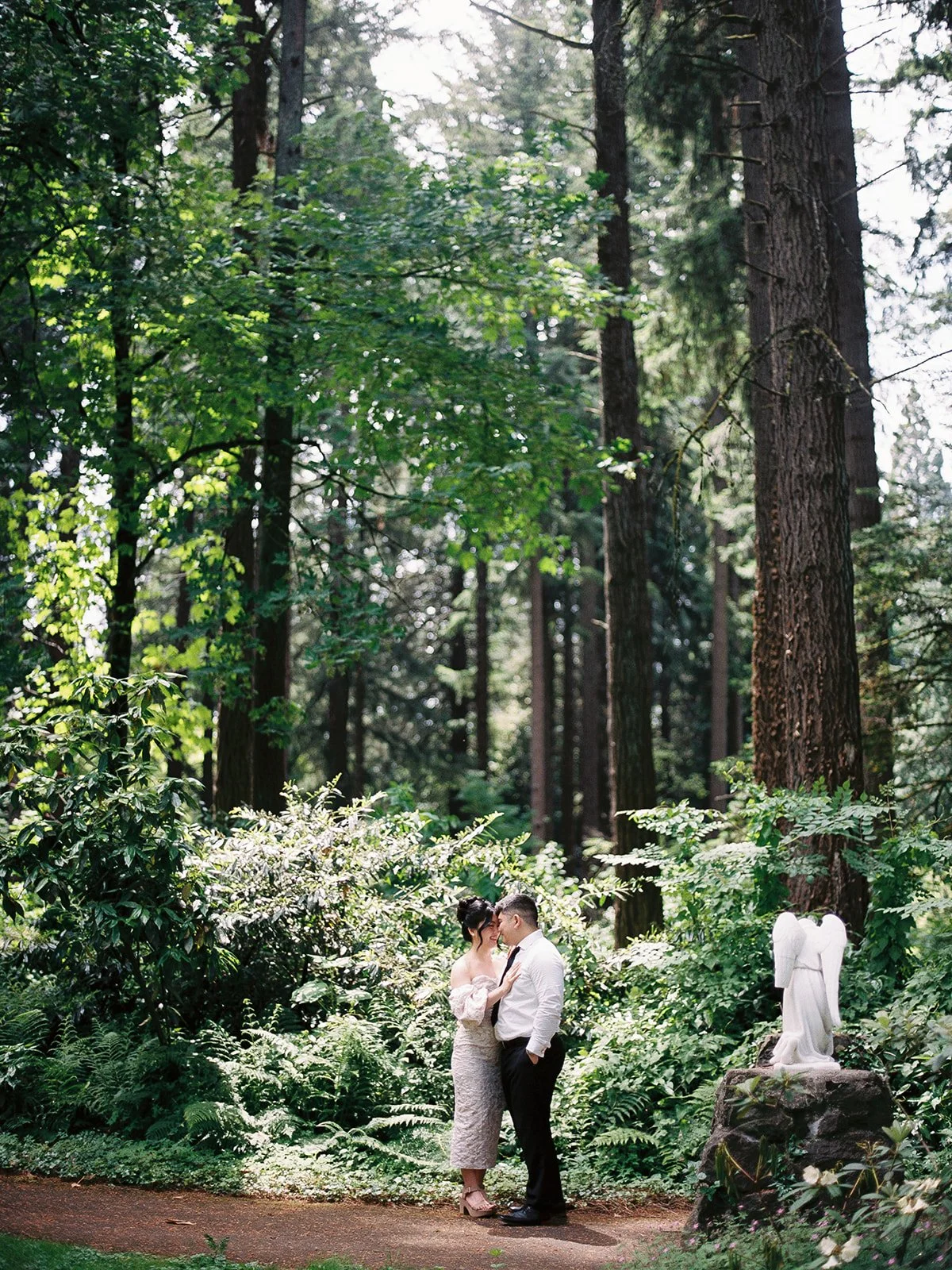 Bride and groom touching foreheads amid towering fir trees and lush ferns at The Grotto Portland, a white angel statue visible among the greenery behind them