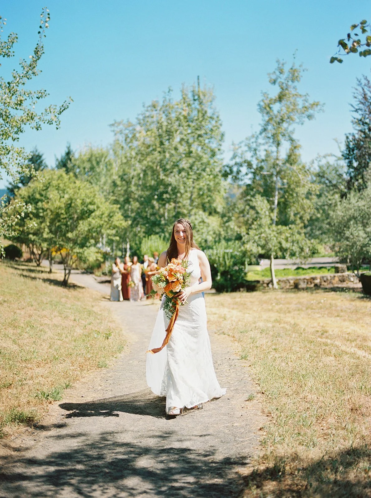 Bride in white dress walking down outdoor path holding bouquet, bridesmaids behind