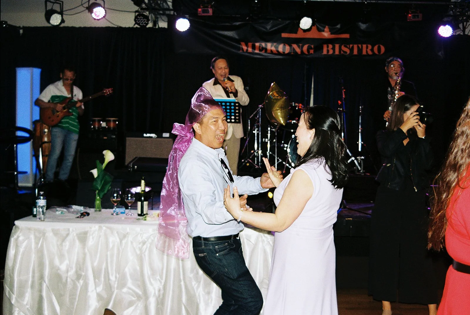 A guest wearing a pink bow on his head dancing enthusiastically with a woman on the Mekong Bistro dance floor, the live band performing behind them