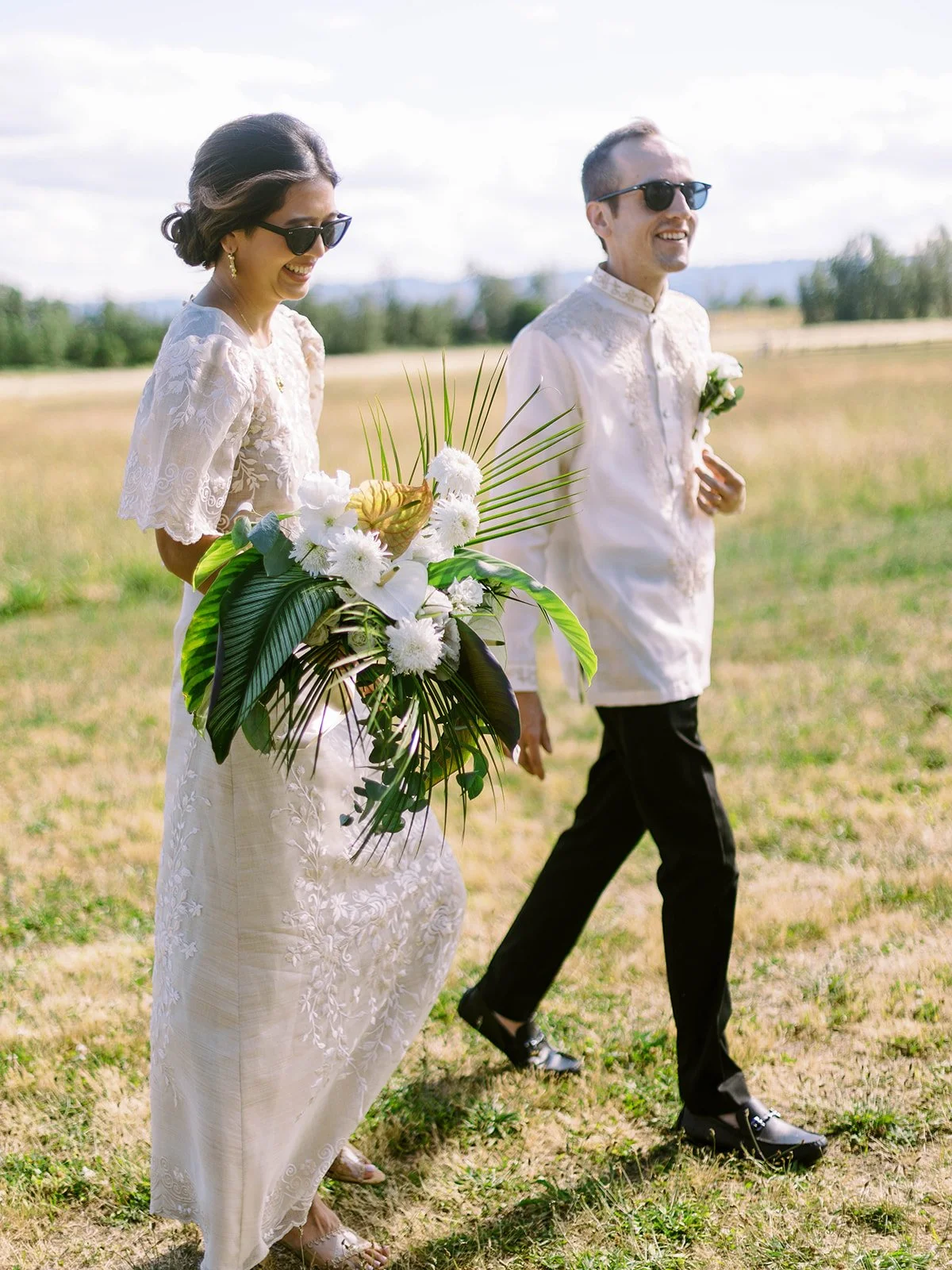 Bride carrying a large tropical bouquet walking ahead of the groom through a grassy field, both wearing sunglasses
