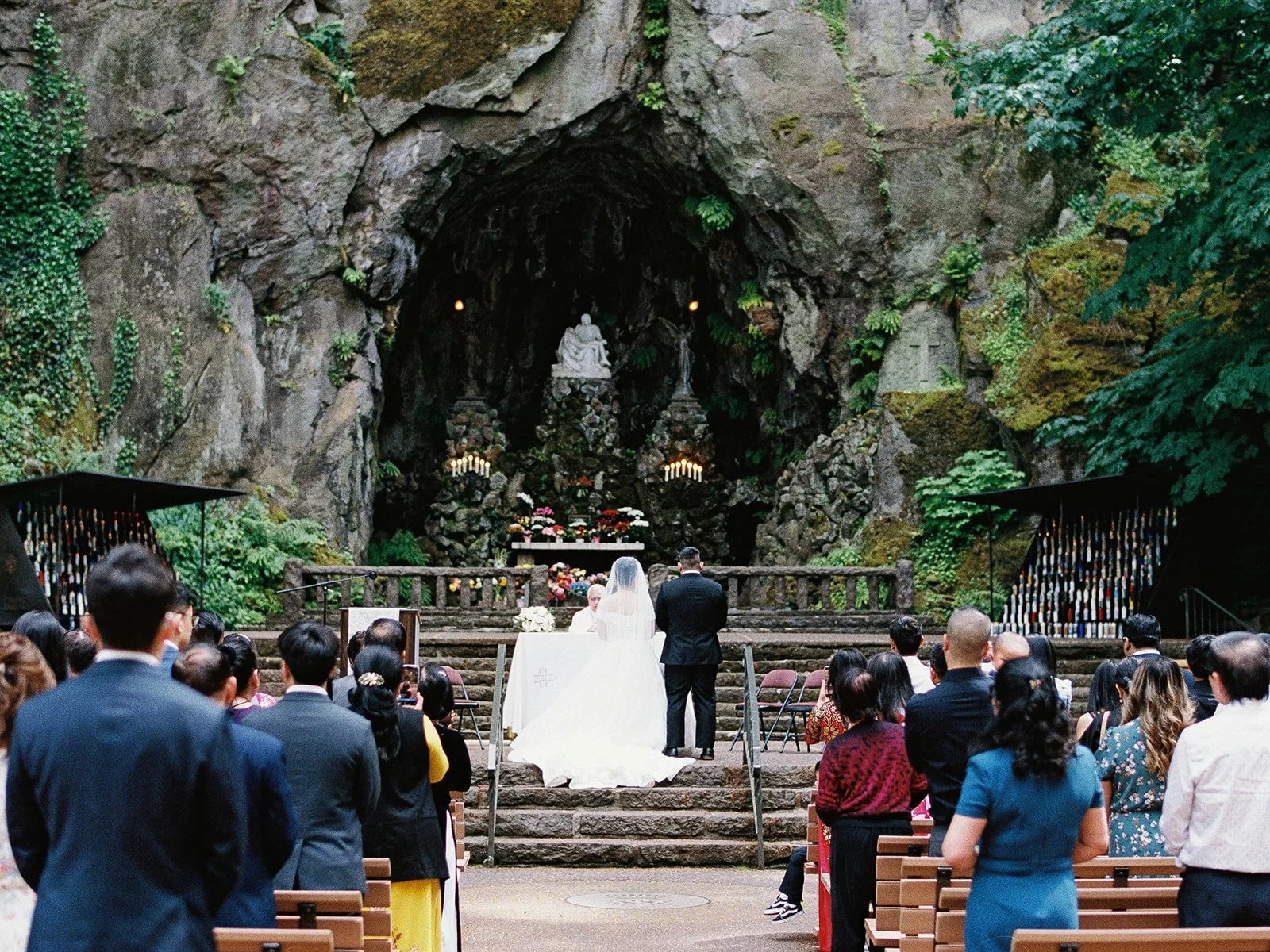 Wide ceremony shot of the bride and groom at the altar before The Grotto's massive basalt cave, the Pietà sculpture and candlelit shrine above, guests seated in wooden benches below