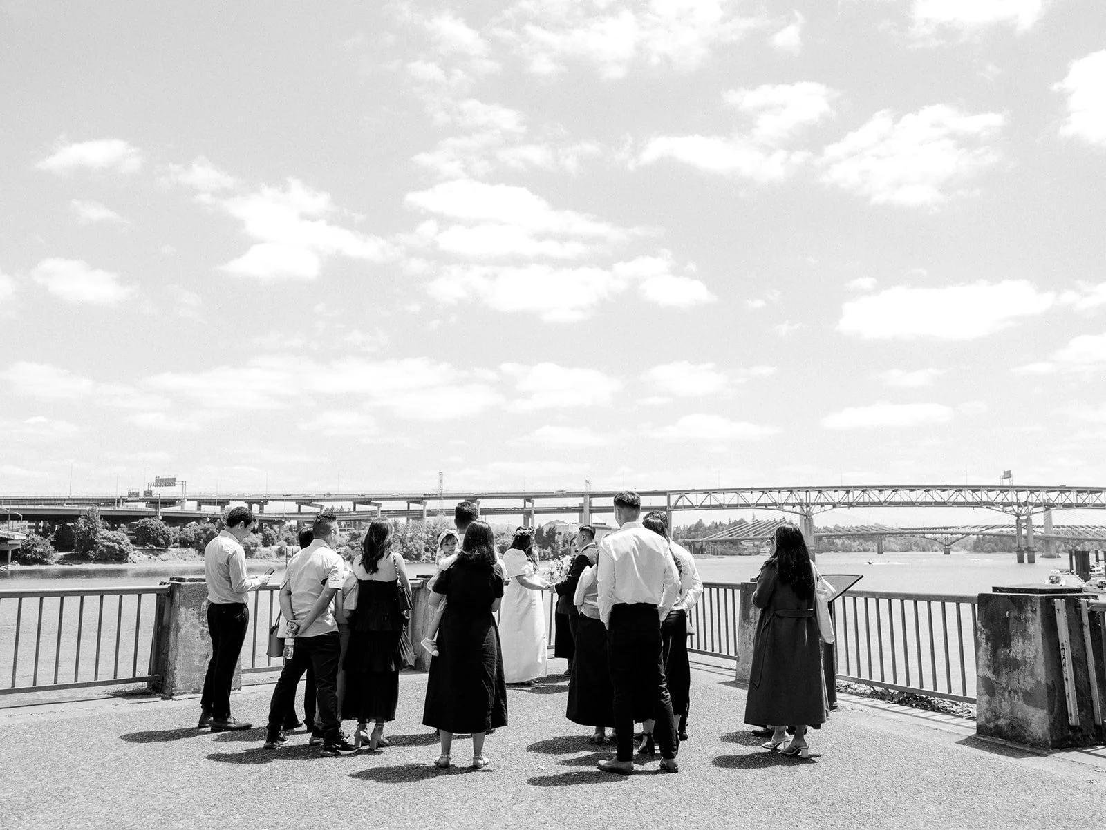 Black and white wide shot of wedding guests gathered around a couple during an outdoor ceremony beside the river.