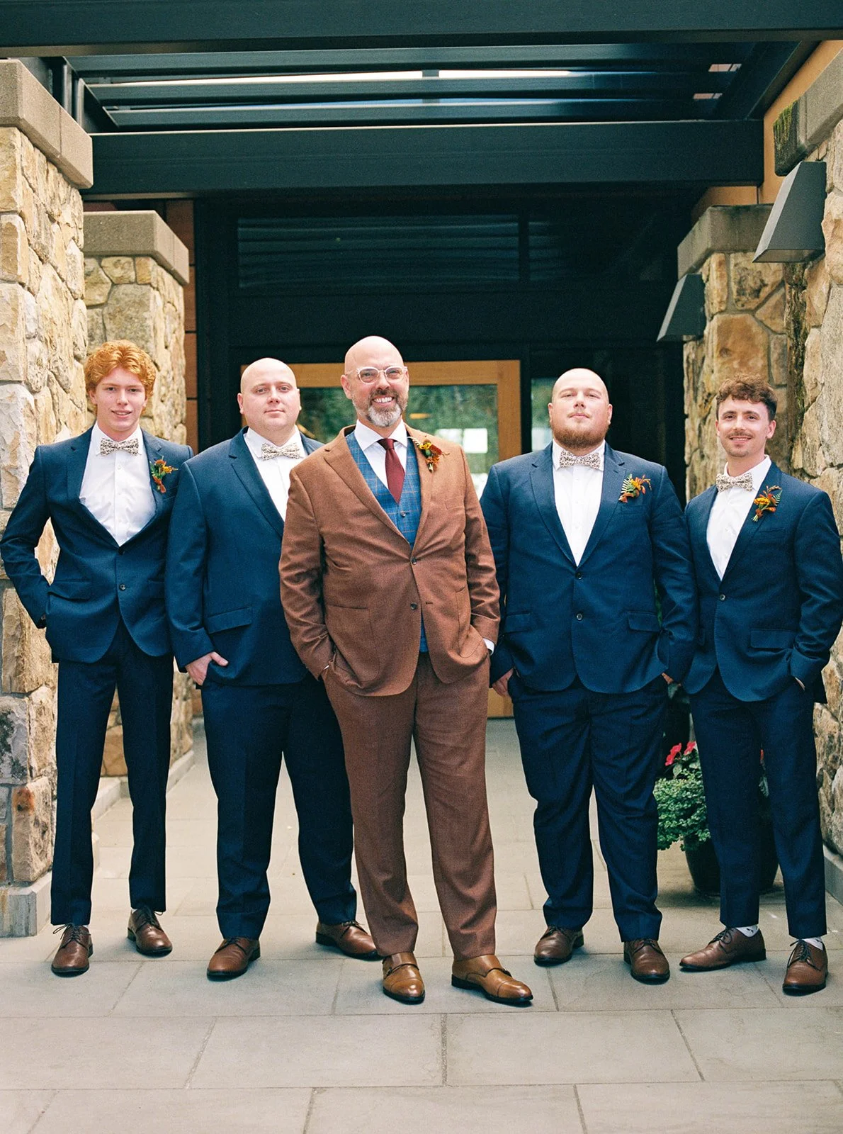 Groom in a brown suit standing with four attendants in navy suits outside a stone building.