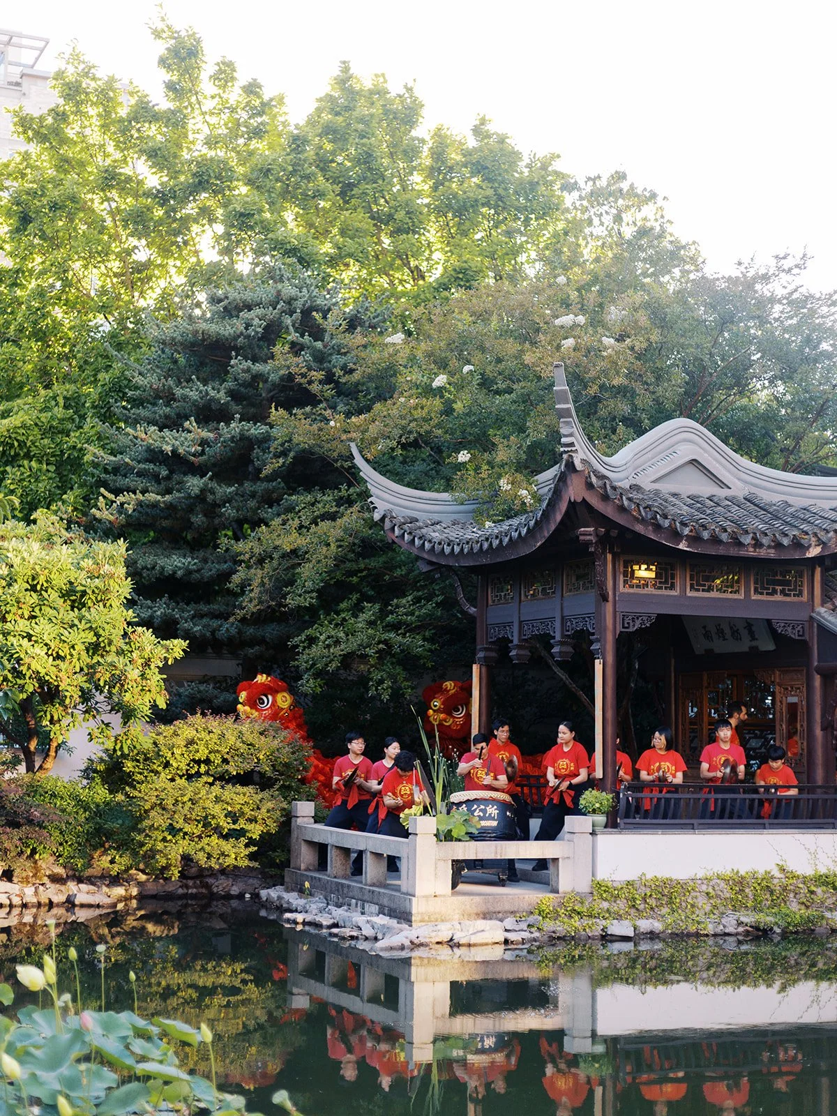 A lion dance troupe in red shirts prepares beside two vibrant red lion dance costumes at the lakeside pavilion, their reflection visible in the still water below.
