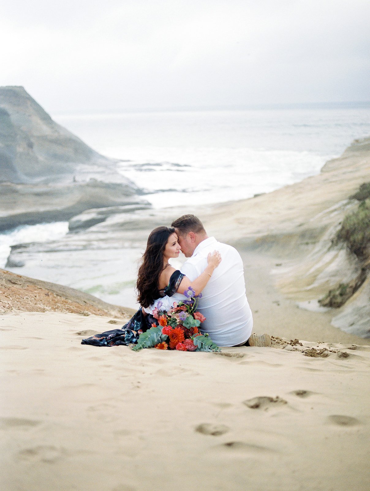 Couple sitting together on sand overlooking Oregon Coast cliffs, bouquet beside them, soft muted tones