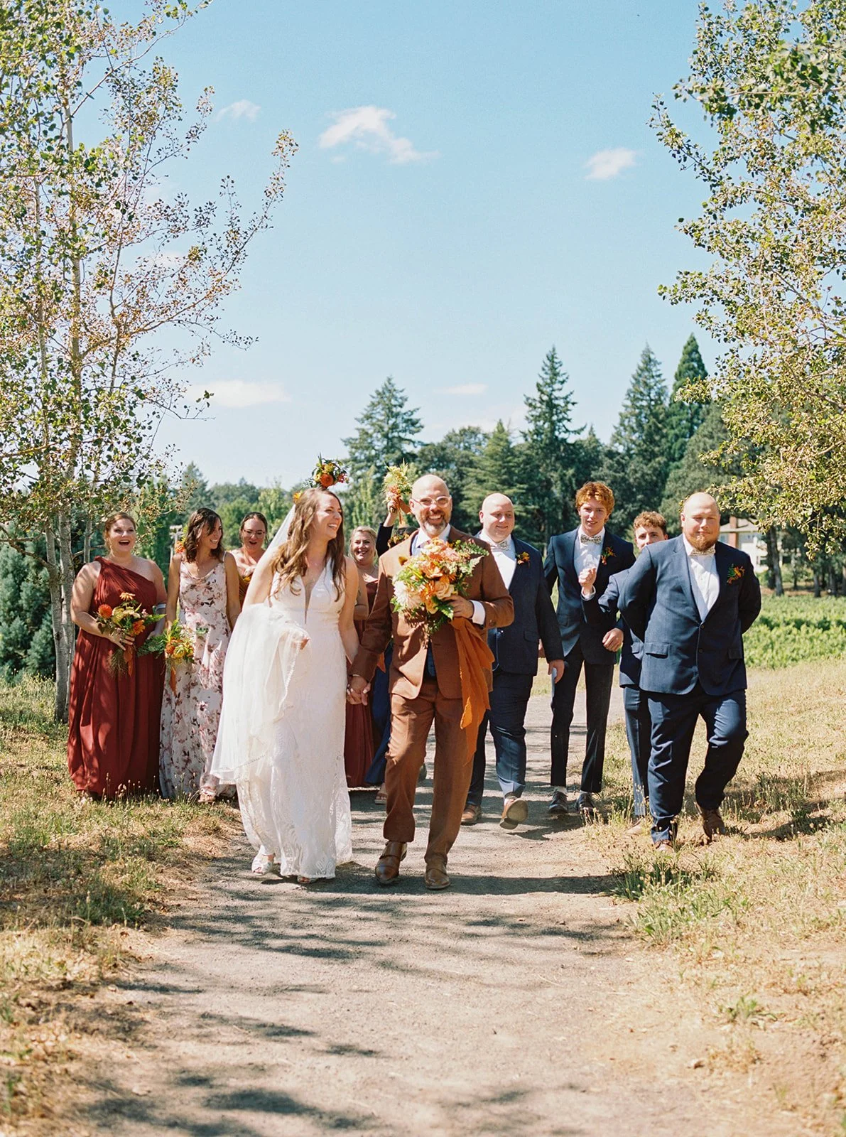 Bride and groom walk down a tree-lined path with wedding party in earthy-toned attire at an outdoor garden wedding.