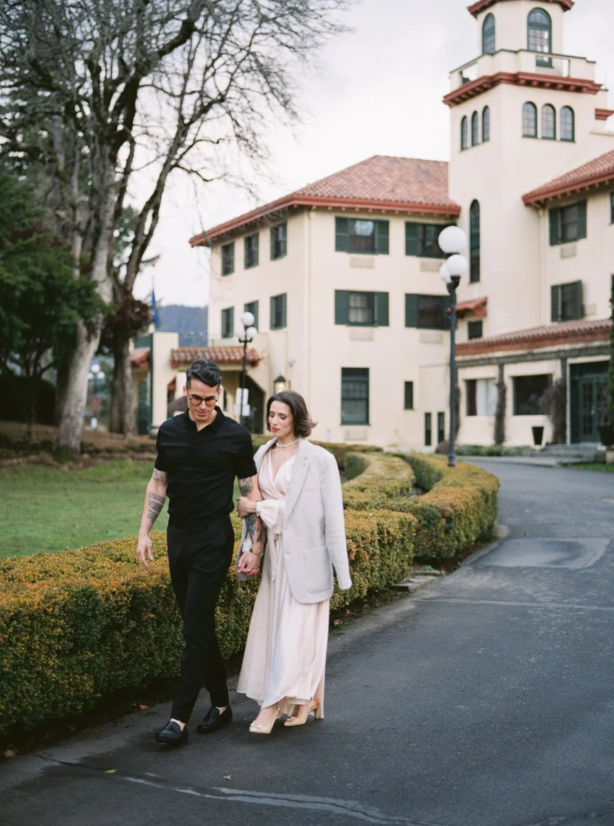 Stylish couple walking along path outside historic building, woman in flowing cream dress and coat, man in black attire