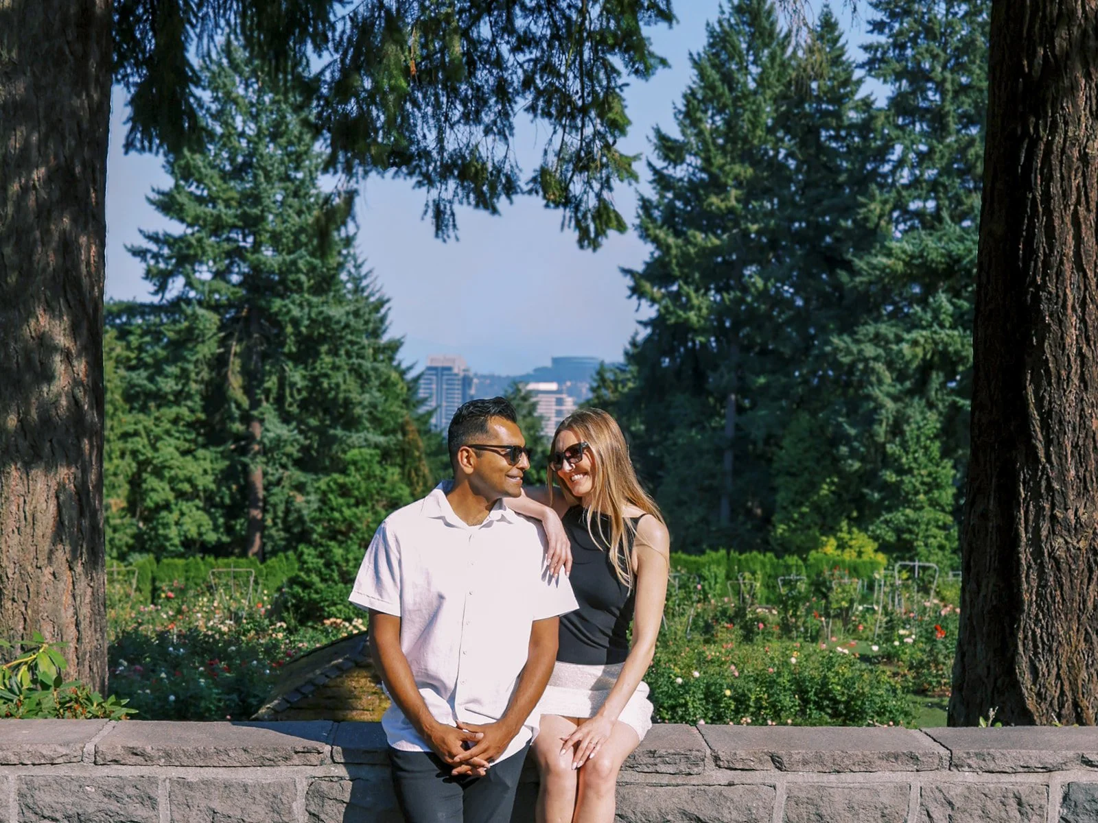 A smiling couple in sunglasses seated on a stone wall framed by tall trees, with the Portland skyline visible behind them.