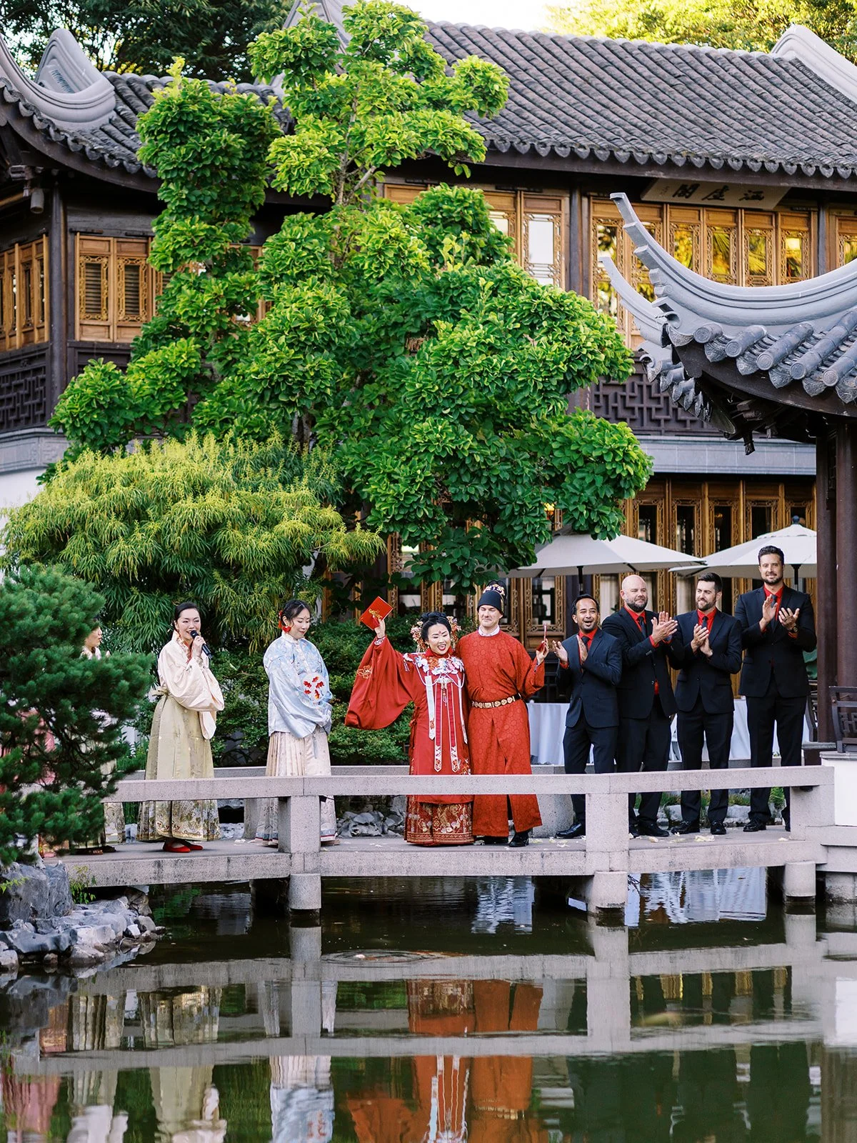 The couple in red traditional robes raise their hands in celebration on the stone bridge, flanked by attendants in hanfu and groomsmen in black suits, the garden reflected below.