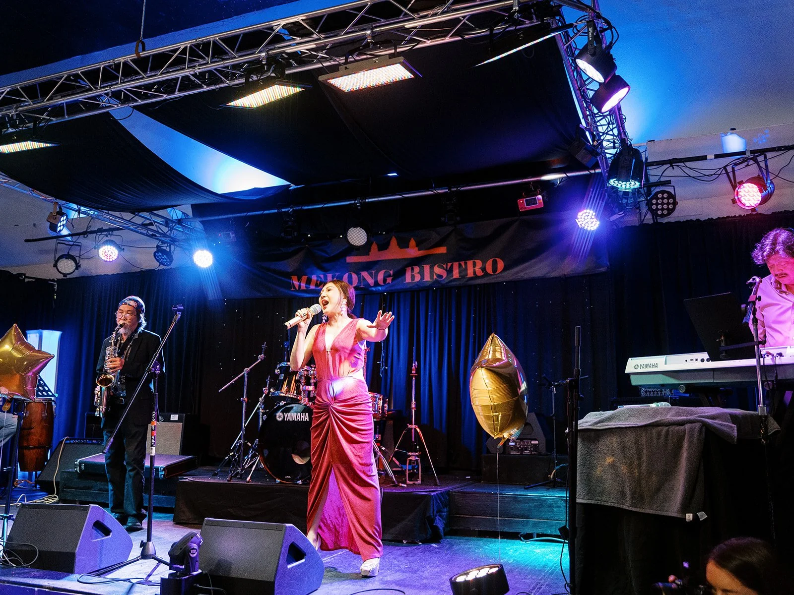 A female vocalist in a pink-red gown performing with arms outstretched on the Mekong Bistro stage, backed by a full live band including a saxophonist and keyboardist