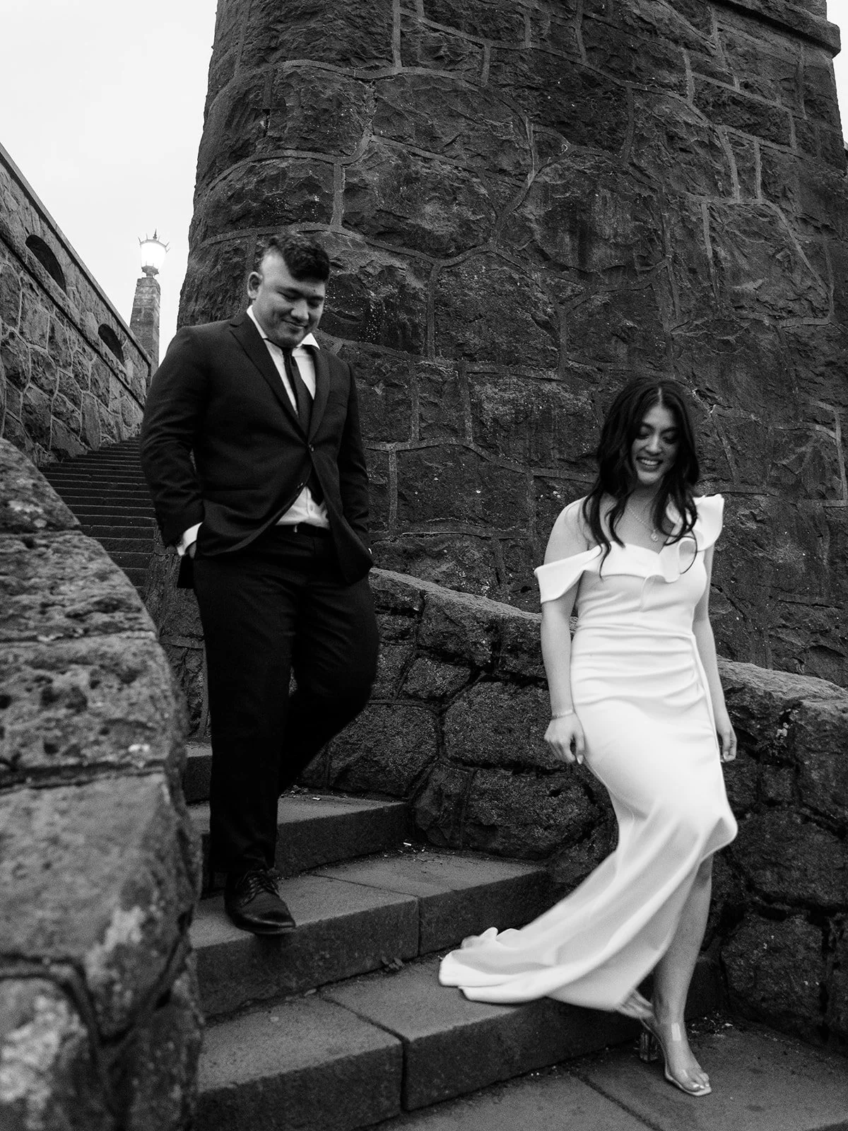 Black and white photo of the bride and groom navigating the stone steps at The Grotto's upper overlook, the massive basalt pillar and lamp post behind them, both looking down and smiling