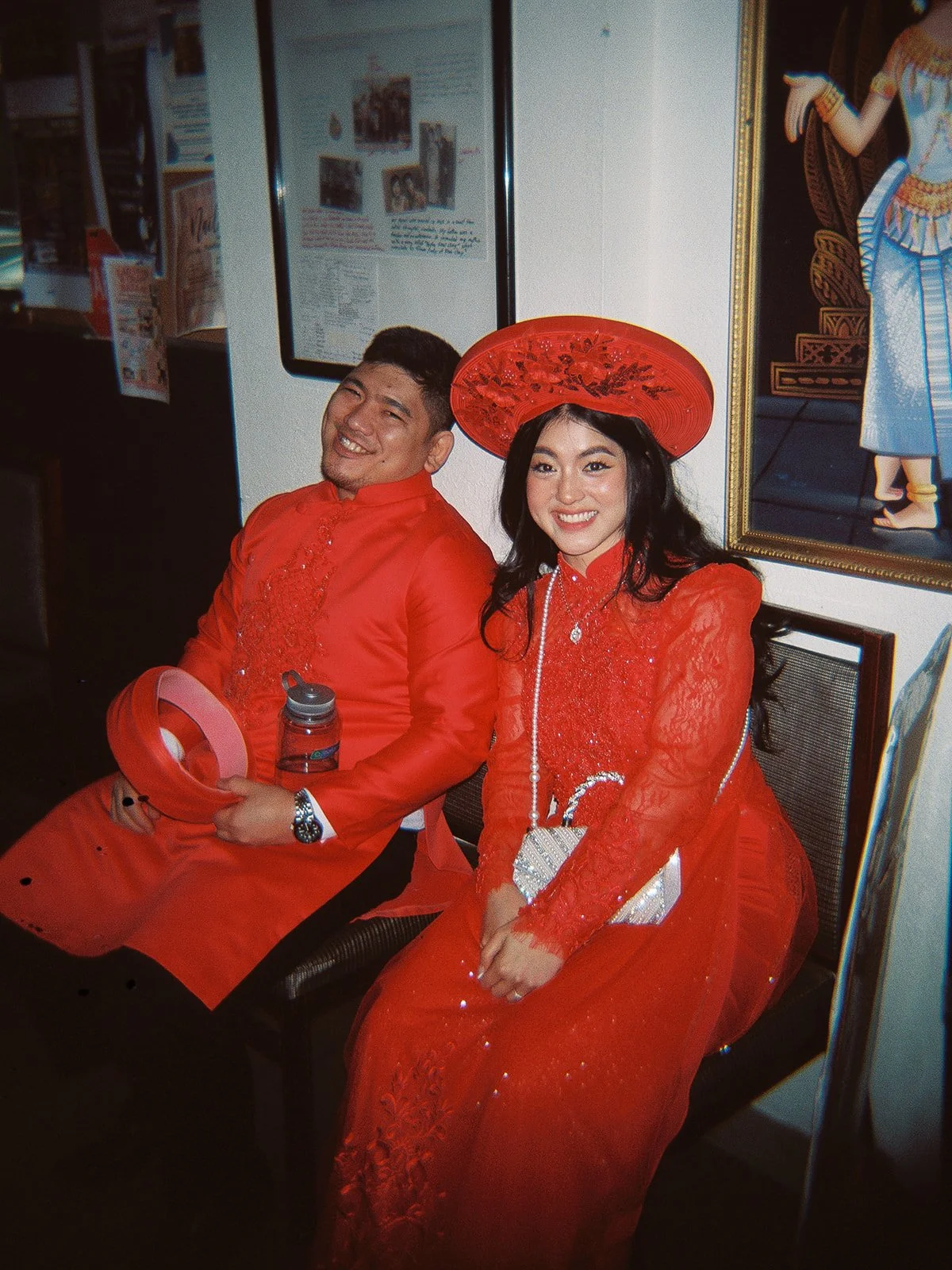 Candid shot of the bride and groom seated together backstage in their red Vietnamese áo dài, both smiling, traditional artwork and framed photos on the wall behind them