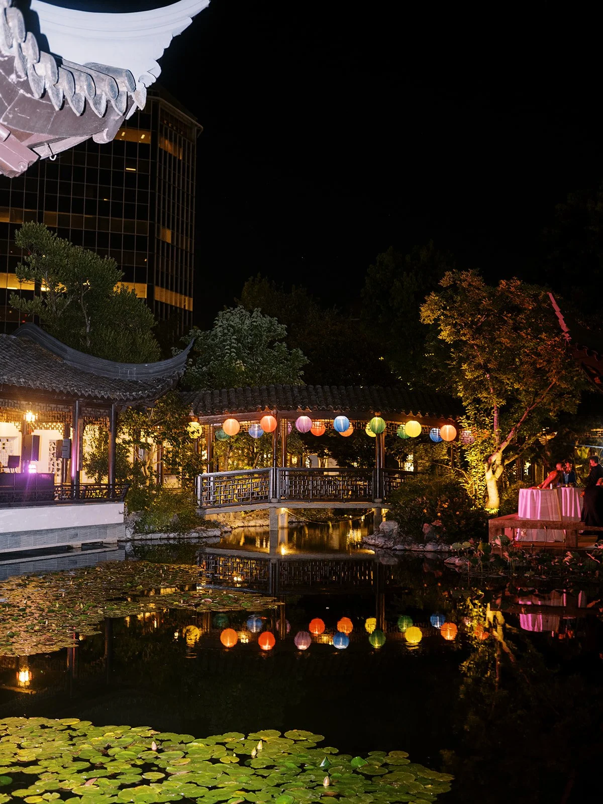 A string of colorful paper lanterns illuminates the covered bridge at Lan Su Chinese Garden at night, their reflections glowing in the dark lily-pad-covered lake below.