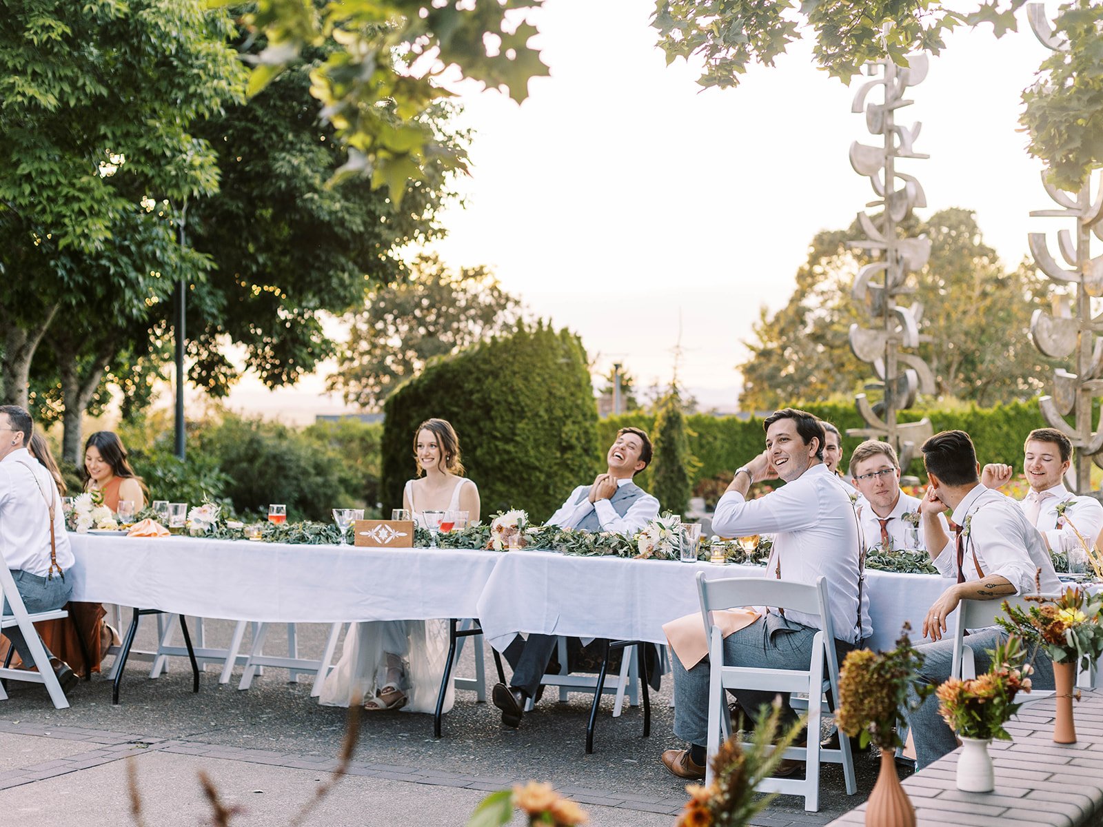 Bride, groom and wedding party seated together at a long reception table during an outdoor garden dinner.