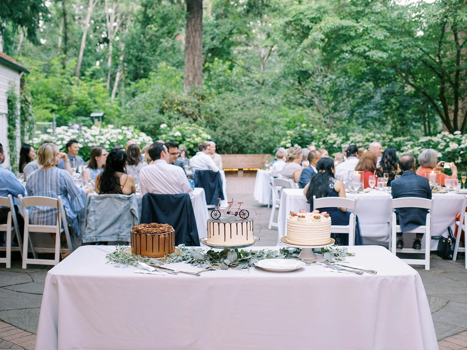 Three wedding cakes displayed on a greenery-covered table during the garden reception.