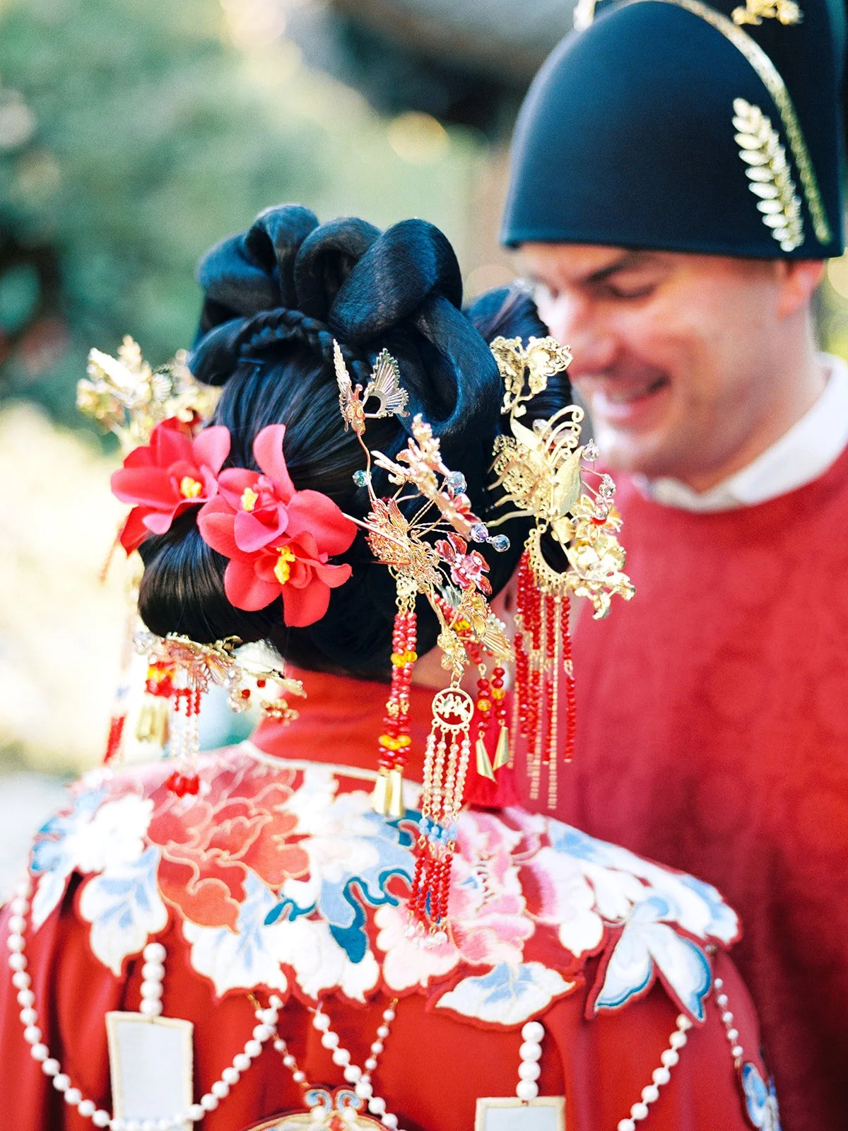 Close-up detail of the bride's elaborate traditional hair ornaments — red flowers, gold phoenix pins, and dangling crystal tassels — with the groom smiling softly behind her