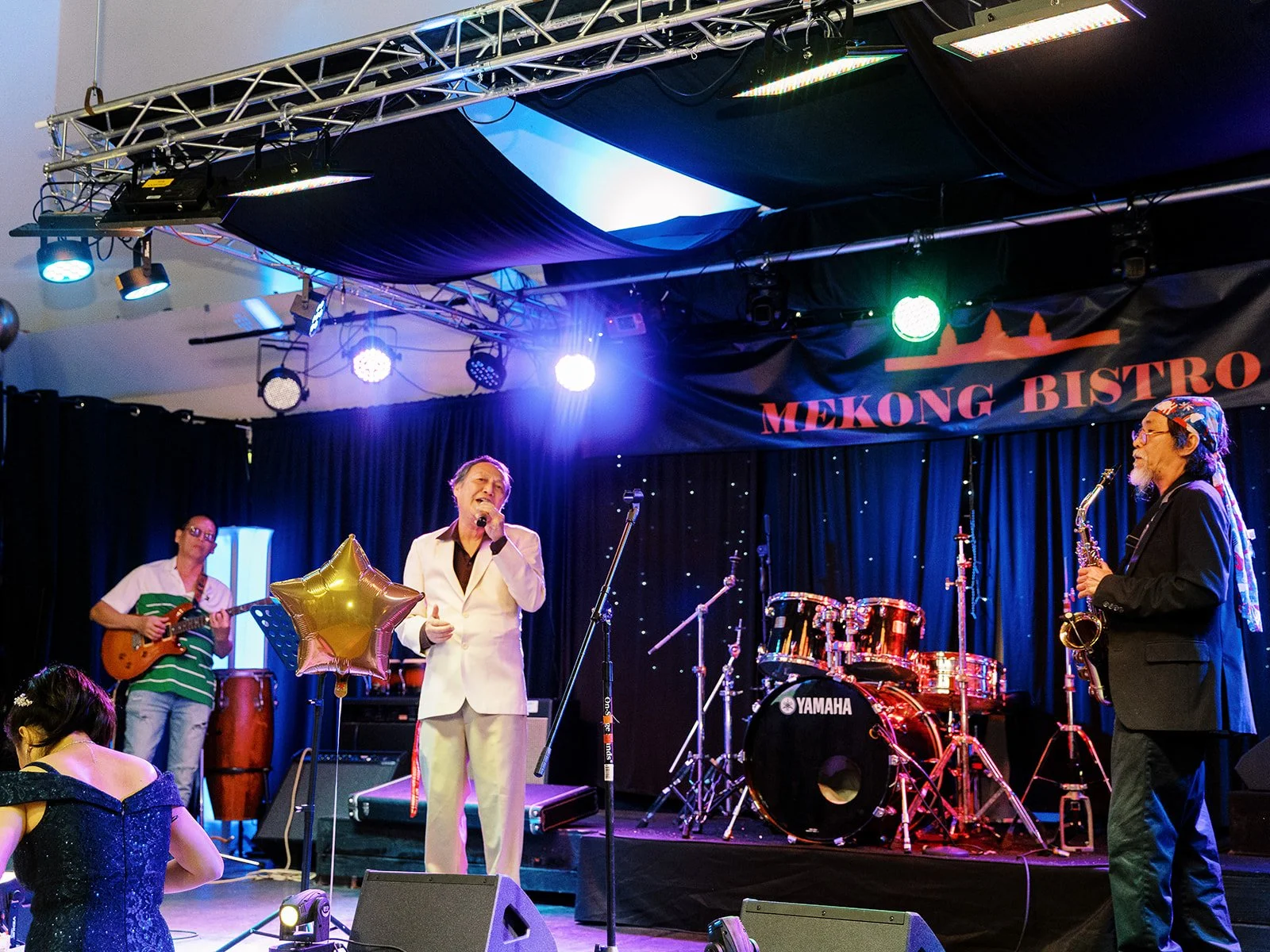 A male singer in a white suit performing on the Mekong Bistro stage flanked by the saxophonist and guitarist, gold star balloons on either side