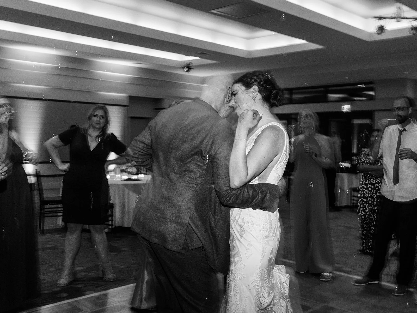 Black and white photo of the couple in an intimate slow dance, the bride laughing softly as guests look on in the background