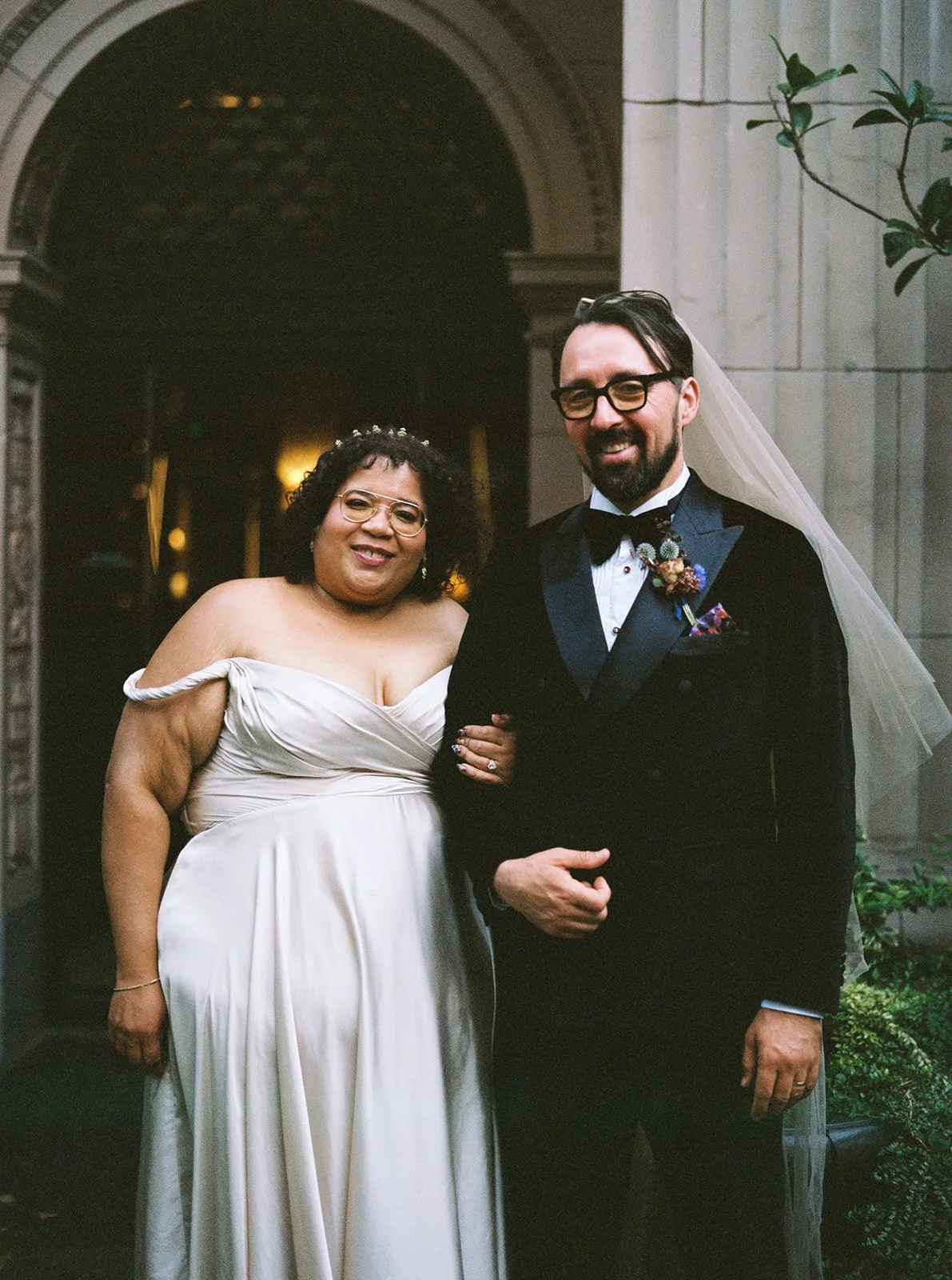Bride and groom posing outside arched doorway, standing arm in arm in front of historic building entrance.