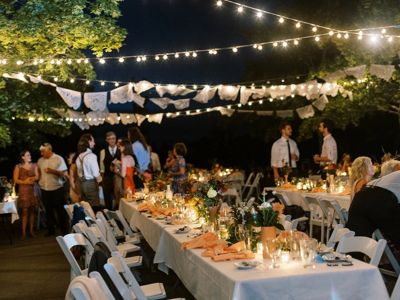 Outdoor wedding reception at night with long tables, candles, and string lights overhead