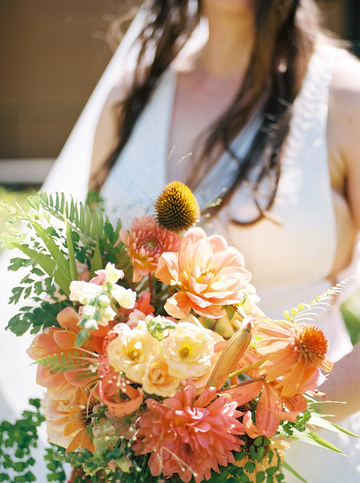 Bride holding a loose orange, peach and cream wedding bouquet in bright outdoor light.