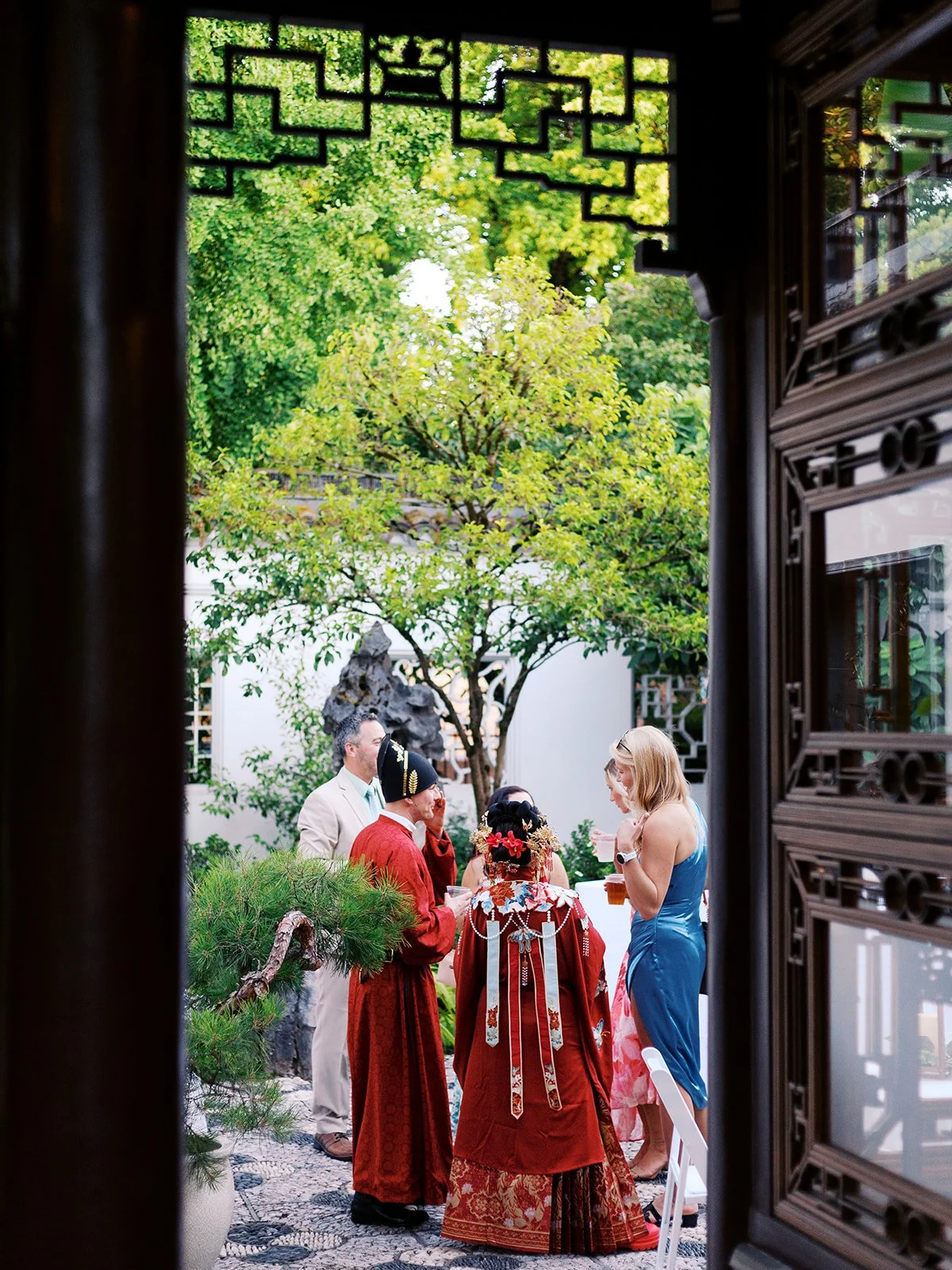 The couple in red wedding robes seen from behind through a latticed moon door, socializing with guests in a lush courtyard.