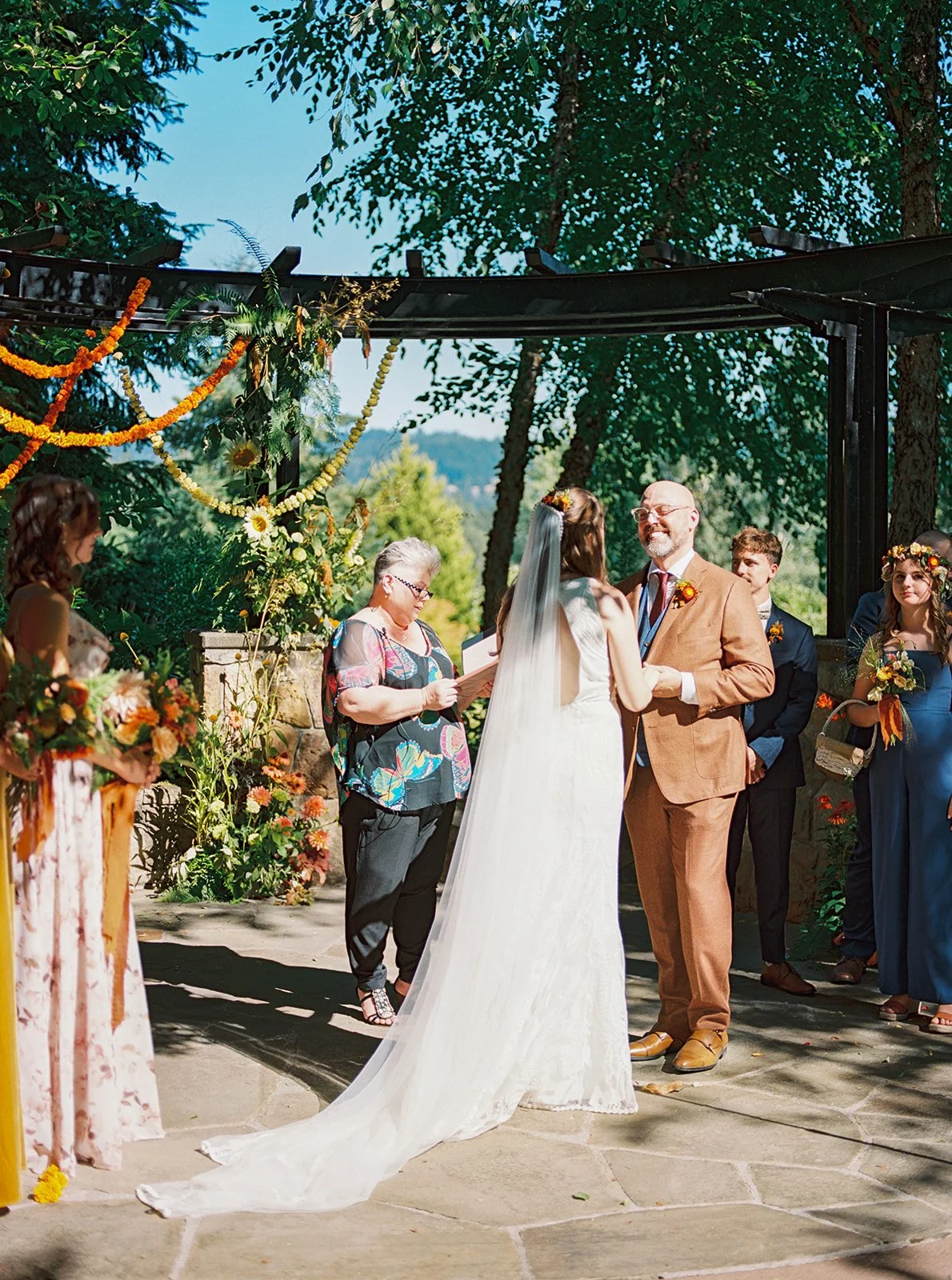 Bride and groom exchange vows under a decorated arbor with floral arrangements and wedding party nearby.