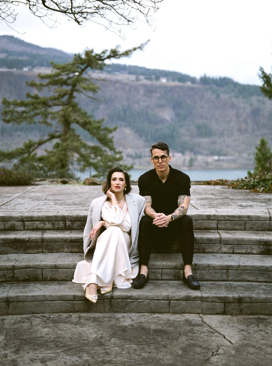  A couple seated on stone steps overlooking the Columbia River Gorge — she in a white dress and blazer, he in all black with tattooed arms.

