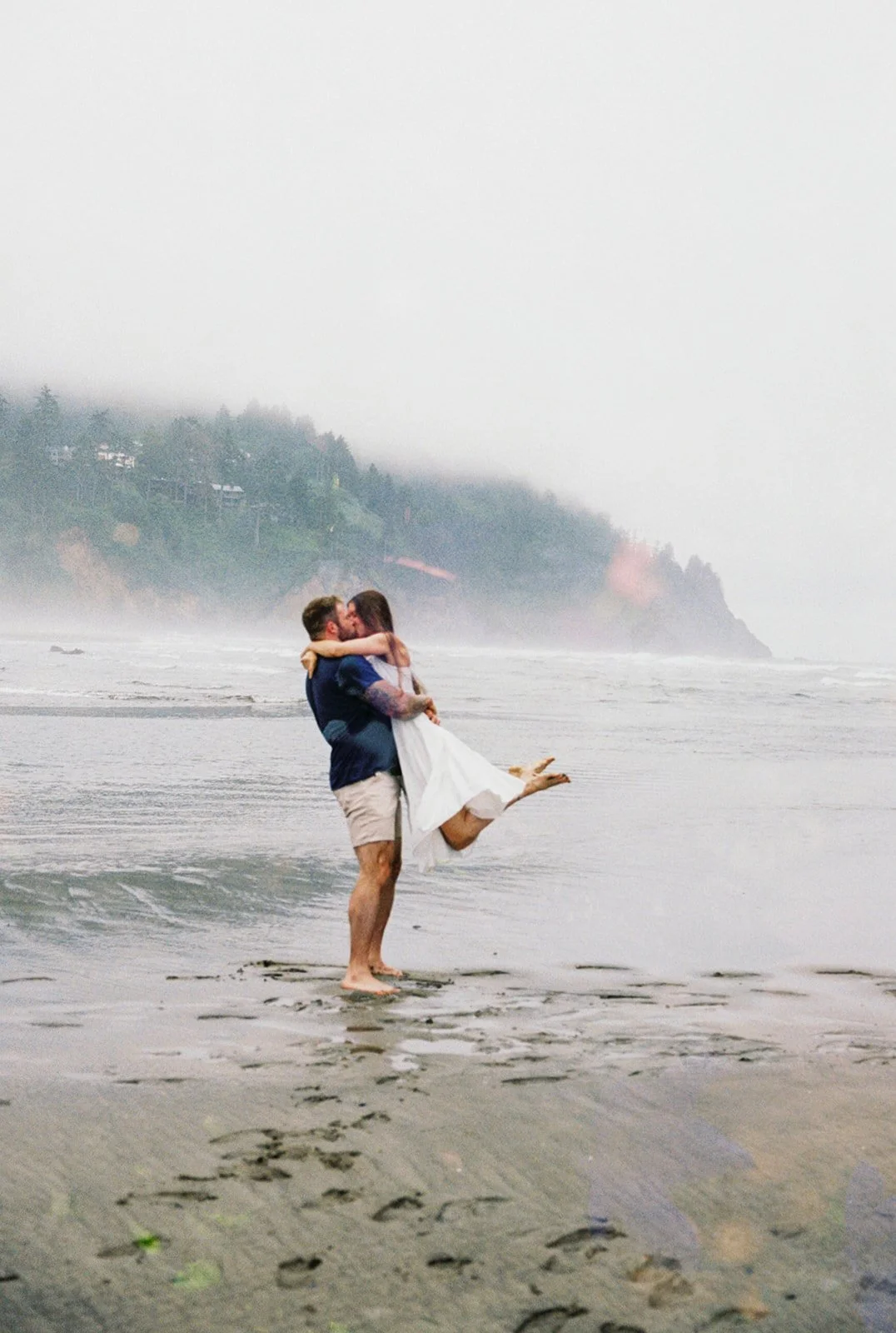 Man lifting woman as they kiss in shallow water on foggy beach with forested cliffs in distance.