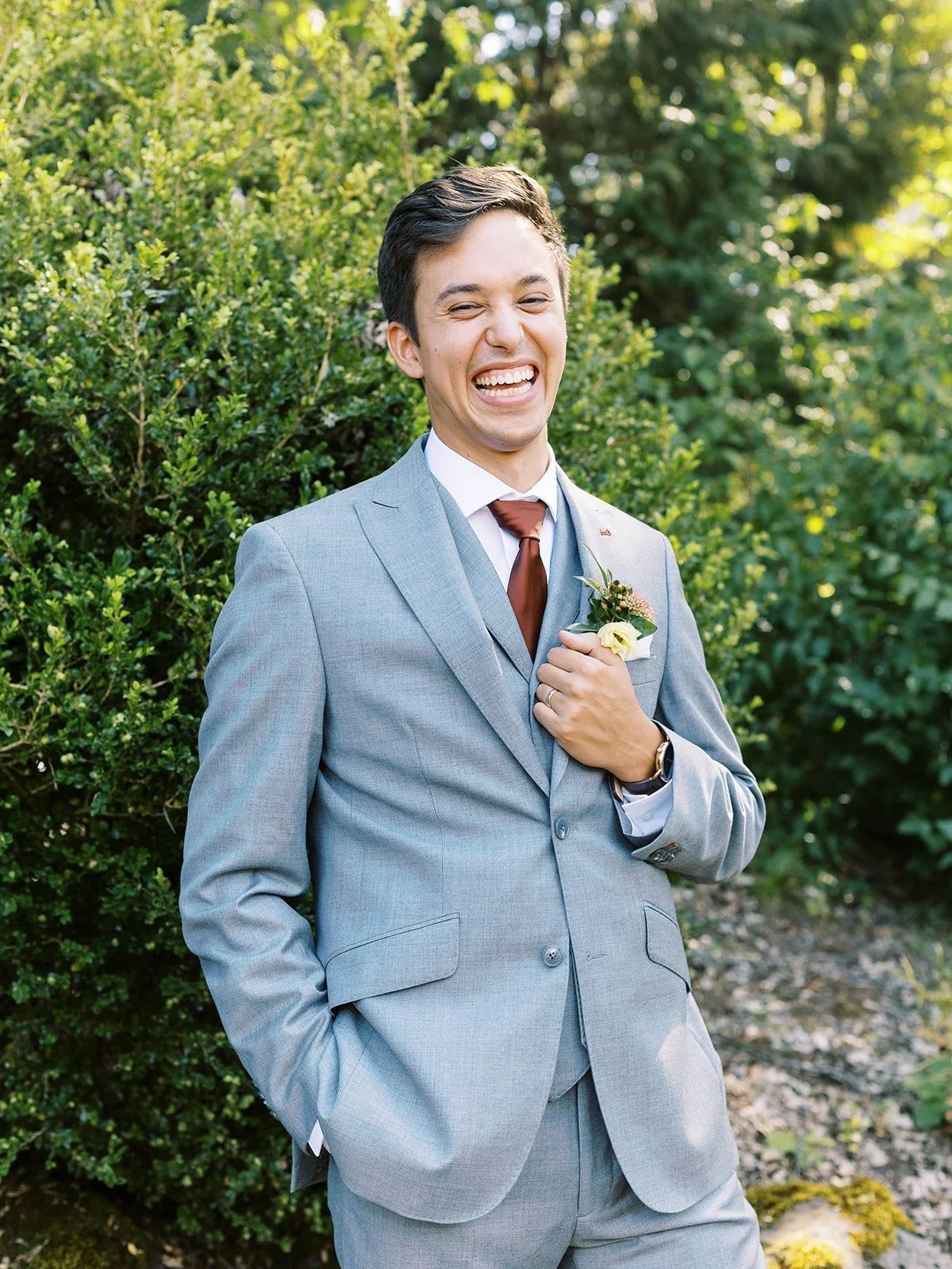 Groom portrait in a gray suit with boutonniere, smiling in front of lush greenery at Oregon Gardens.
