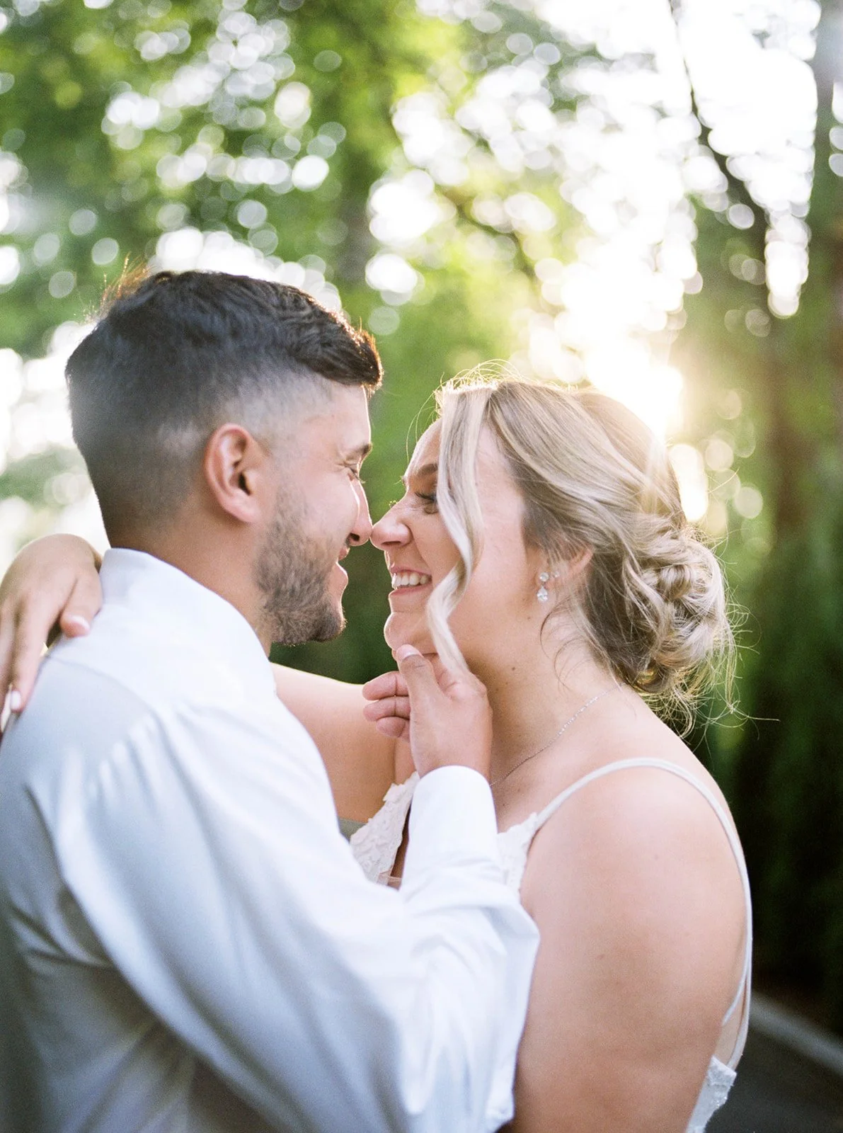 Bride and groom embrace closely outdoors at sunset, smiling with sunlit trees blurred in the background.