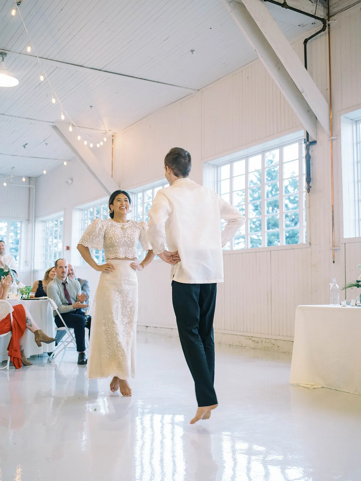 Bride smiling with hands on hips while the barefoot groom hops toward her during their playful first dance inside the Pearson Air Museum hangar
