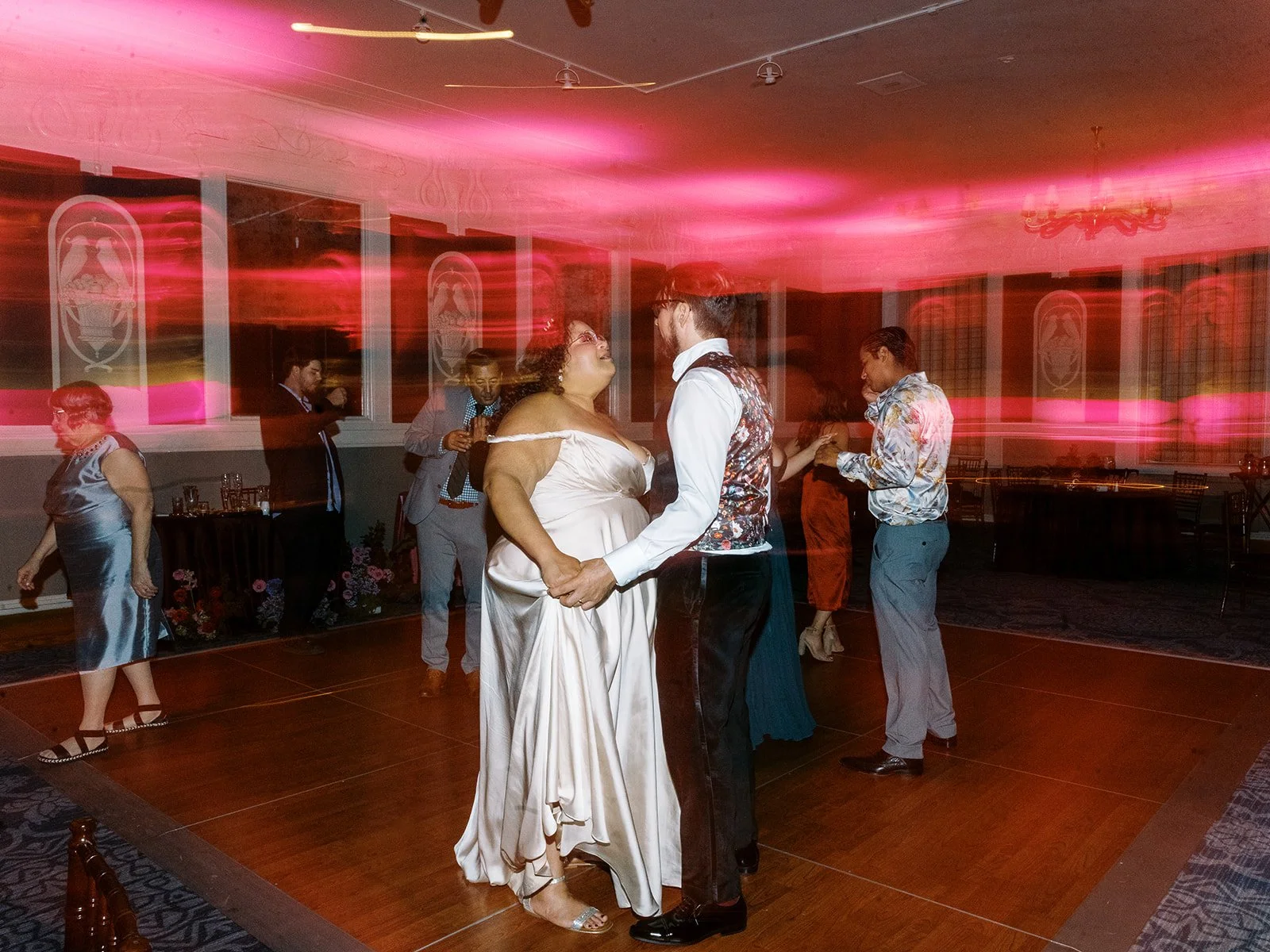 Bride and groom dancing together on dimly lit dance floor with pink light trails and guests nearby.