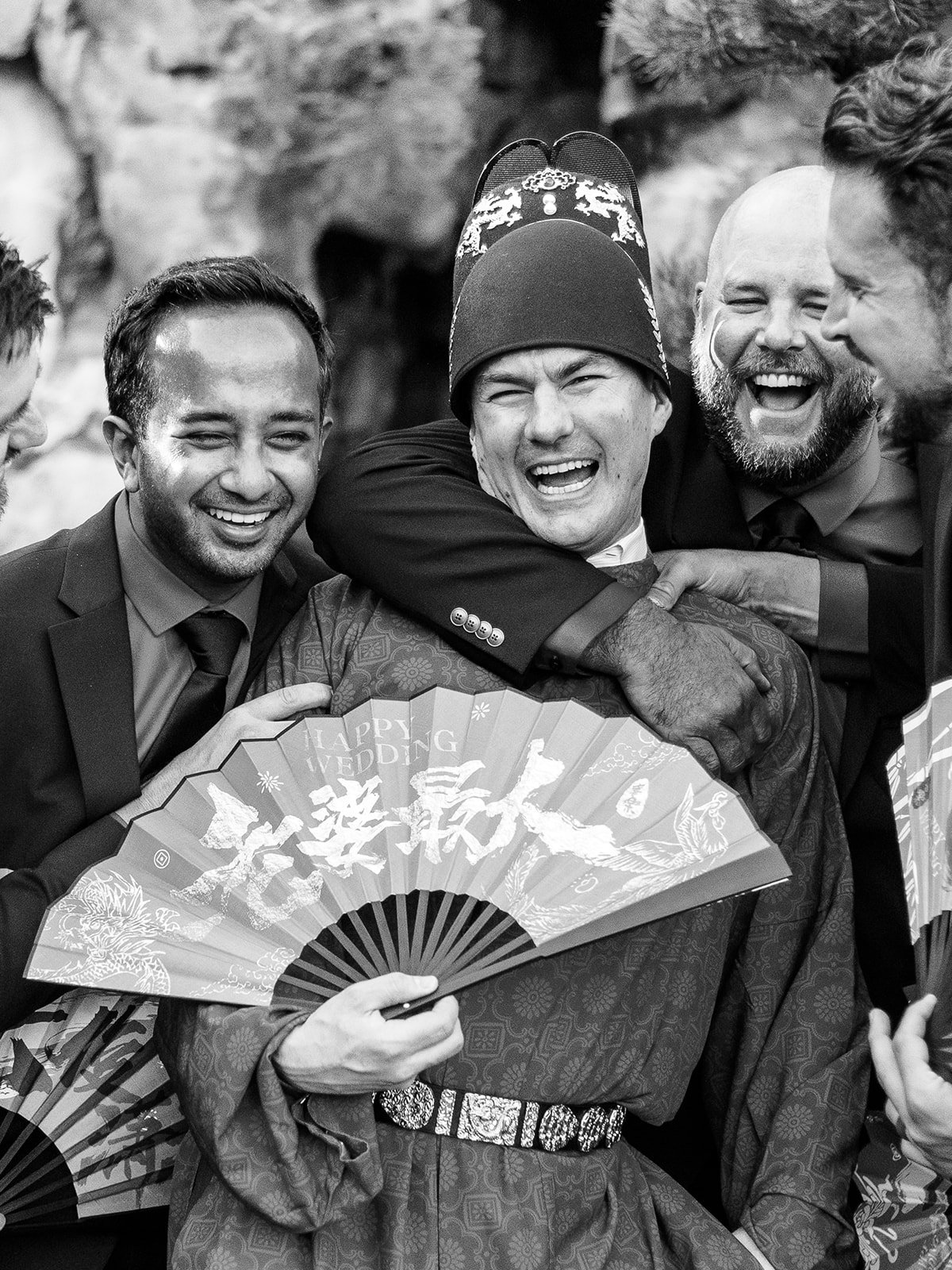 Black and white close-up of the groom in traditional red Chinese wedding robe and ceremonial hat laughing joyfully with groomsmen holding red fans