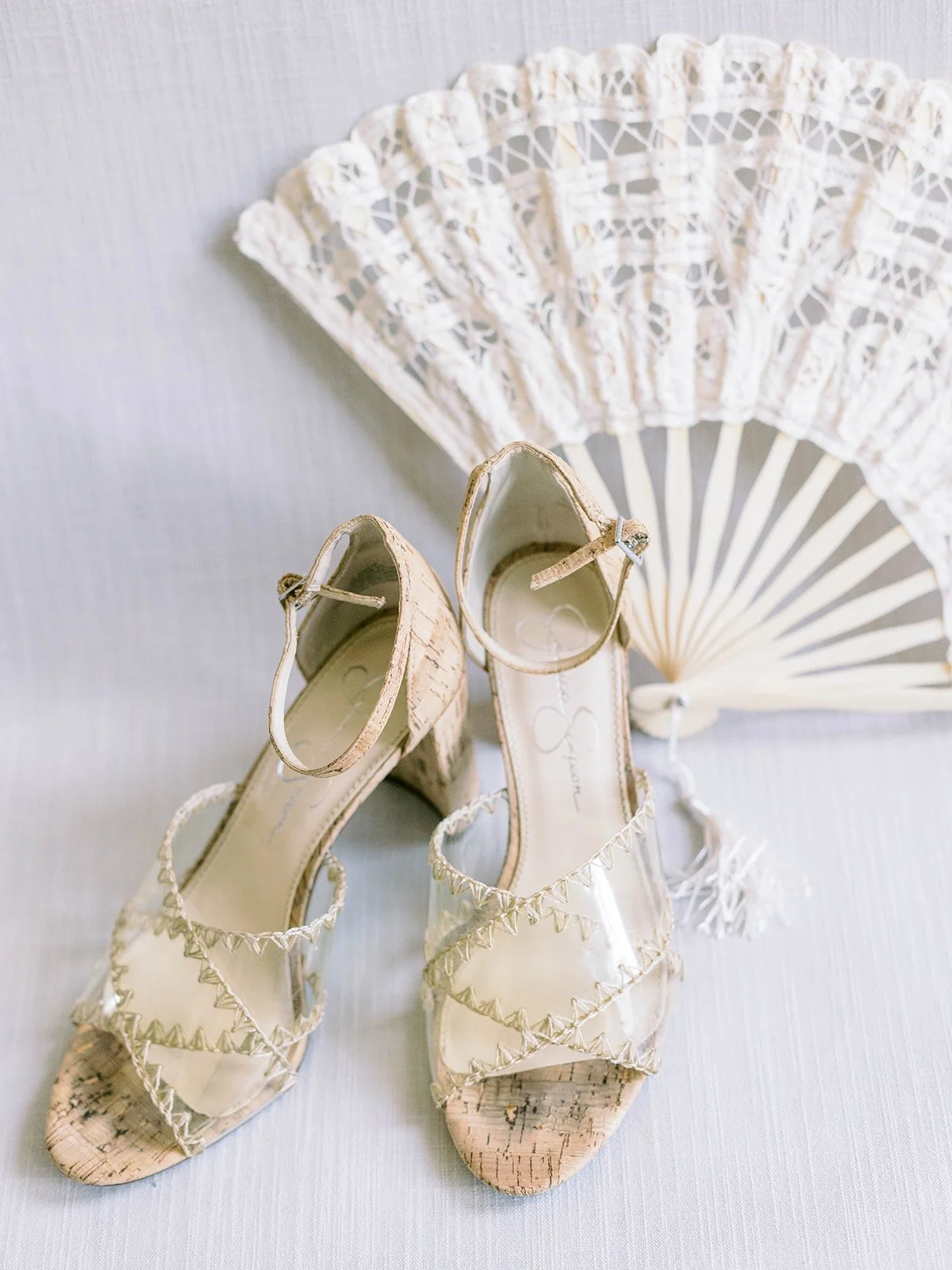 Cork-heeled Jessica Simpson sandals with clear straps and zigzag stitching displayed against a white lace hand fan
