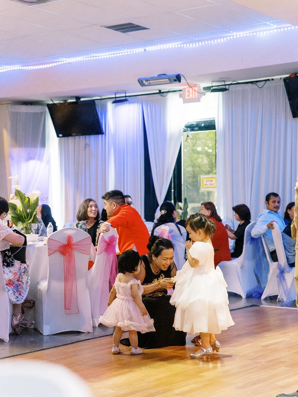 Two young flower girls and a toddler girl on the dance floor with a woman kneeling to tend to them, the Mekong Bistro reception hall glowing behind them